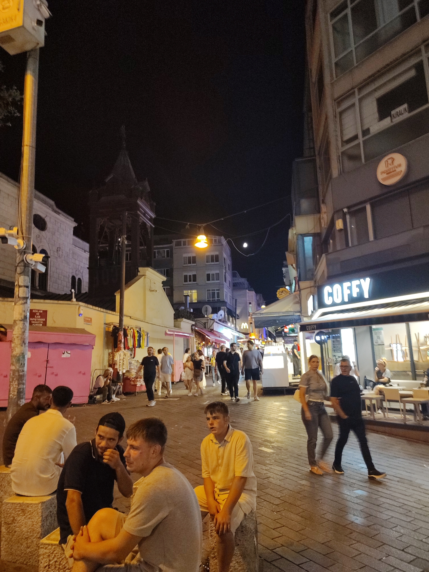 This is a nighttime street scene in Kadıköy, Turkey, featuring a bustling pedestrian area with shops and people. On the left, a multi-story building with white facade and arched windows stands next to a darker, older structure with a pointed roof, possibly a church or mosque. A lamppost with a yellow light illuminates the scene. Pink stalls with hanging clothes are visible along the sidewalk.
In the foreground, several men are seated on stone blocks. Two men in the center are engaged in conversation, with one gesturing. Further right, a young man in a yellow shirt sits alone. Across the street, a cafe with outdoor seating is lit up, with patrons visible inside and at tables. A prominent sign reads "COFFY" in large white letters. People are walking along the cobblestone street, some appearing to be shoppers or diners. Buildings line the right side of the street, with commercial establishments on the ground floor. The overall atmosphere is lively, indicative of an active urban nightlife. The presence of CCTV cameras on a pole on the left suggests a monitored public space.