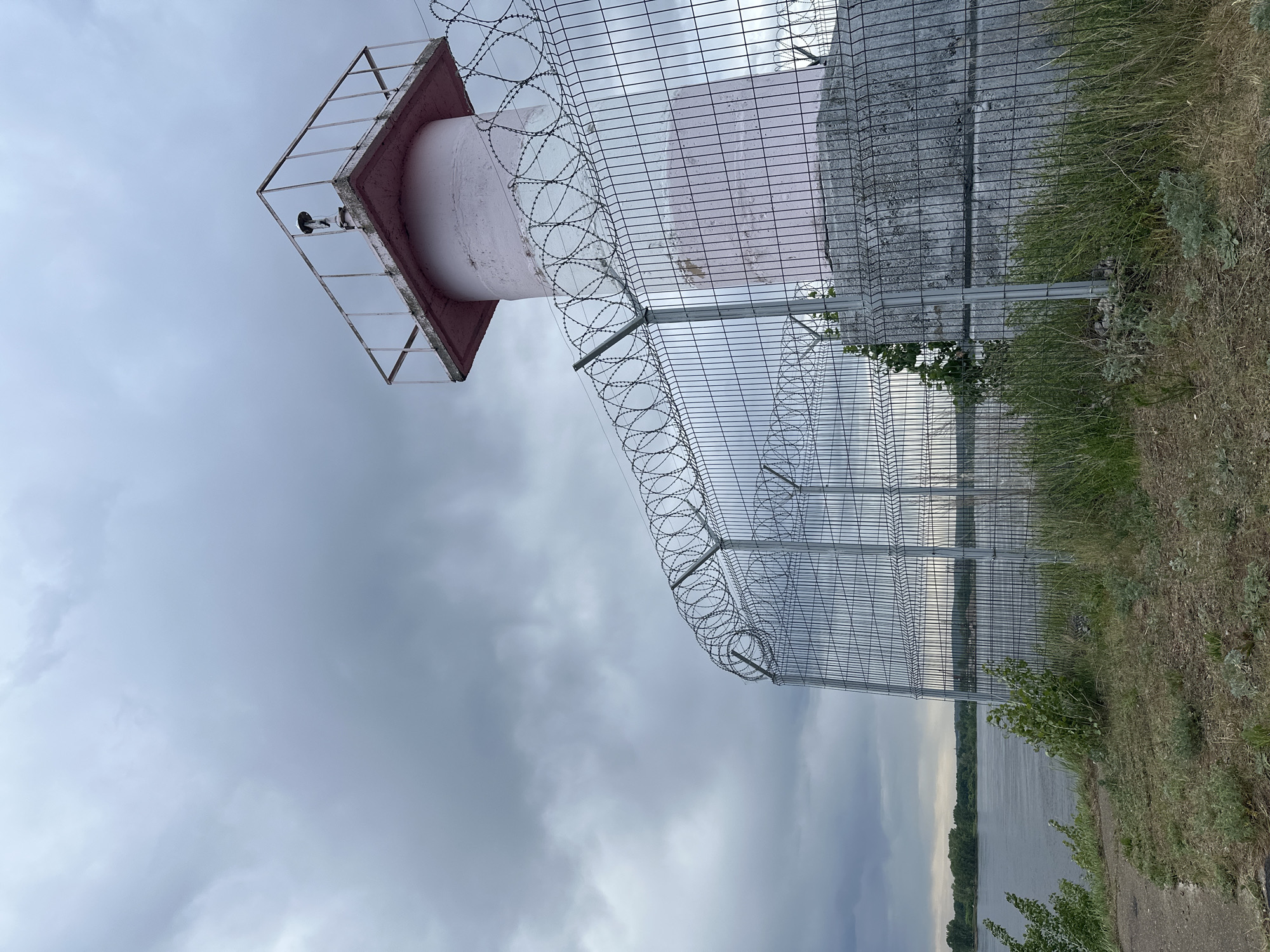 A partially obscured, low-angle shot reveals a tall cylindrical structure with a square, red-trimmed top, secured within layers of wire fencing, including razor wire. The location appears to be the Votkinsk hydroelectric power plant in the city of Chaikovsky, Russia, based on the presence of the characteristic signal lights and the proximity to the water reservoir of the Votkinsk hydroelectric power plant. The metal mesh fence runs along a grassy embankment. In the background, a body of water reflects the overcast sky. There are no people visible in the frame.