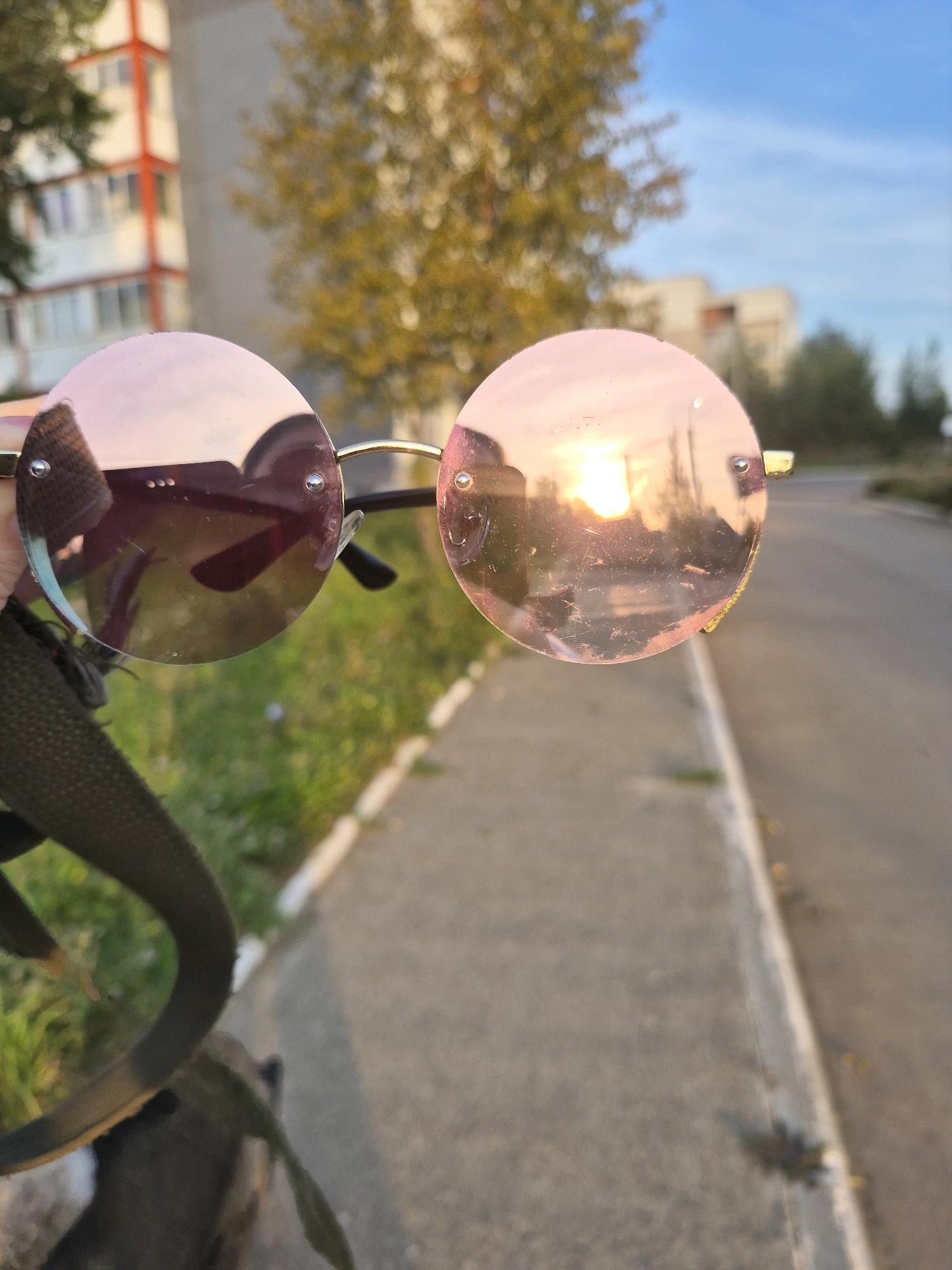 The image shows a close-up of round, rose-tinted sunglasses being held by a person's hand. The sunglasses are reflecting the setting sun and the surrounding environment, which appears to be an outdoor urban setting. In the background, there is a tall, light-colored building with orange accents, and trees with yellowing leaves. A paved sidewalk runs alongside a road. The overall impression is a casual snapshot taken during golden hour. The location is identified as Chaikovsky, Russia, though this detail is not visually discernible from the image itself.