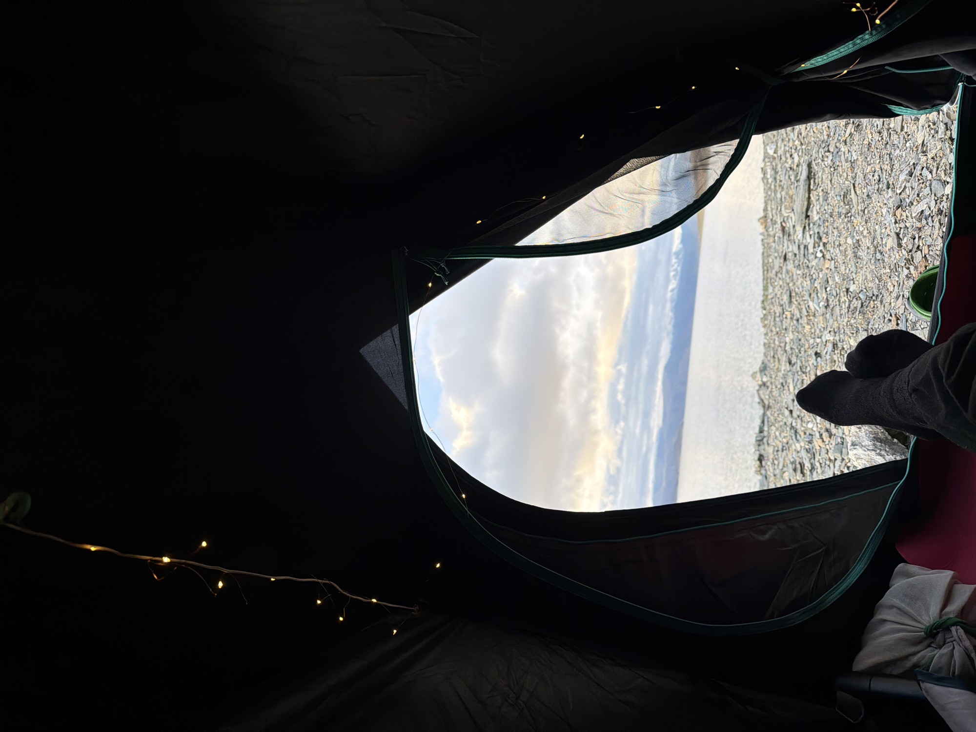 The image is taken from inside a dark tent, looking outwards through an open flap at a body of water and a rocky shore. A string of small, yellow fairy lights is draped along the inside of the tent flap and across the roof. In the foreground, the lower legs and feet of a person wearing dark socks and dark pants are visible, with one foot resting on a sleeping mat. The ground outside the tent is covered in small, grey stones and pebbles. Beyond the shore, a vast expanse of water stretches towards a distant mountainous landscape under a cloudy sky with hints of sunset. The scene evokes a sense of outdoor camping.