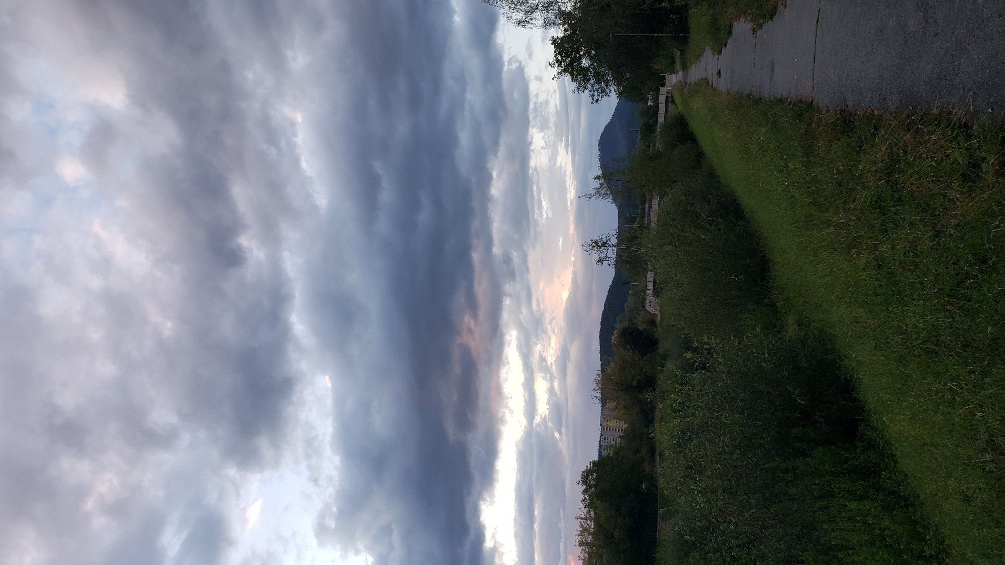 The image depicts a path running alongside a grassy embankment, leading towards a town in the distance. The sky is filled with dramatic, dark clouds, with hints of lighter sky and faint pink hues suggesting either dawn or dusk. Trees and vegetation line the path and the embankment. In the background, there are buildings and hills. The scene appears to be a natural or semi-urban landscape.