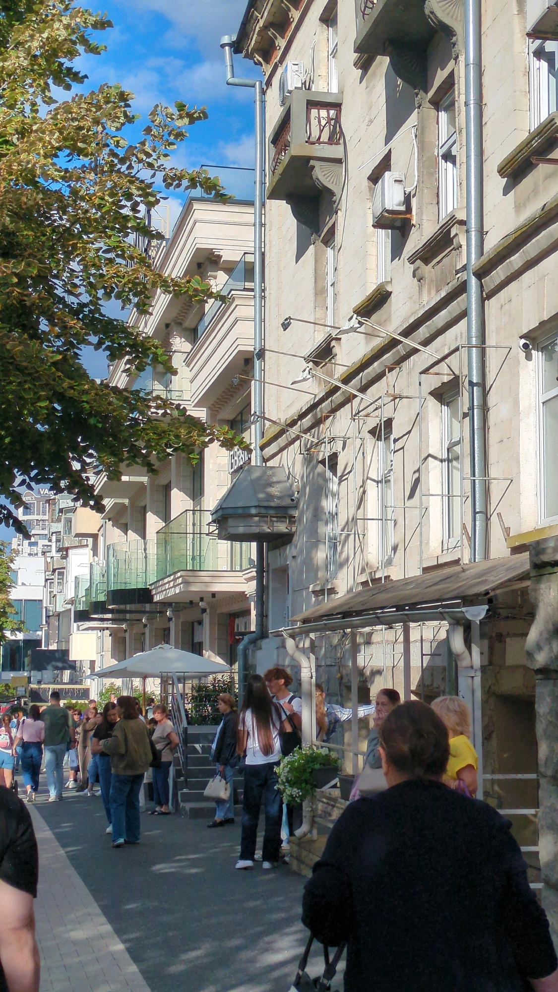 The image captures a street scene in Chișinău, Moldova, on a bright, sunny day with a blue sky and scattered clouds. The perspective is from street level, looking towards a multi-story building with a light-colored stone facade. The building features several balconies with glass railings, some with decorative metalwork. Vertical silver pipes run down the side of the building, along with what appear to be air conditioning units and external wiring. Trees with green and yellow leaves partially obscure the upper floors of the building.
In the foreground and mid-ground, several people are walking and standing on the sidewalk and street. They are dressed in casual summer clothing, including jeans, t-shirts, and jackets. Some individuals are carrying bags. One person in the foreground is seen from behind, wearing a black jacket and carrying a black bag. There's a white umbrella on the left side of the image, suggesting a possible outdoor seating area or market stall. A lush green plant with white flowers is visible on the right side of the image. The ground is paved with cobblestones and asphalt. The overall atmosphere appears to be that of a lively urban environment.