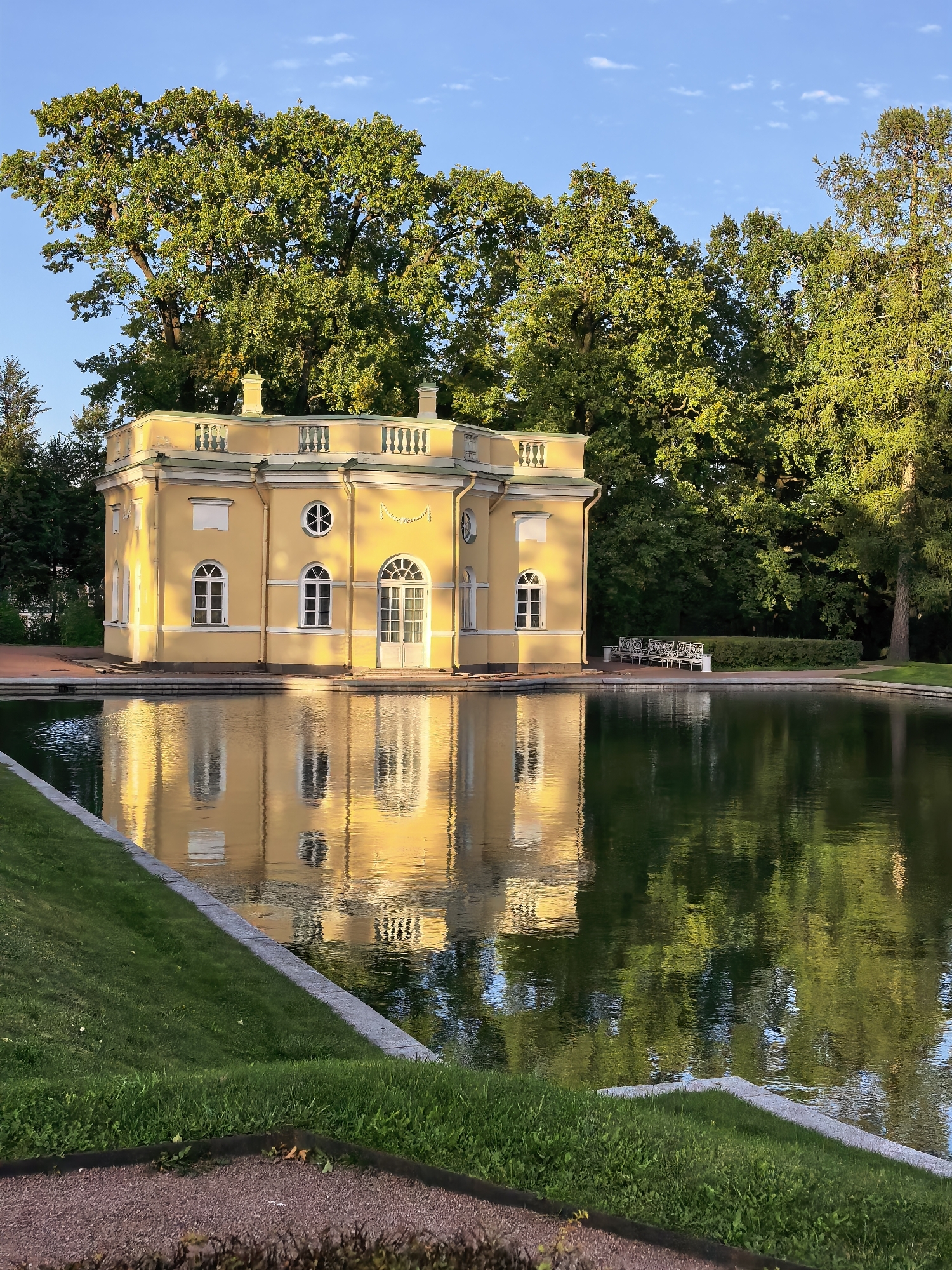 This is a vertical, eye-level, medium shot photograph of a yellow, neoclassical building situated on the bank of a pond in the city of Pushkin, Russia. The building, which appears to be a pavilion or folly, has a symmetrical facade with arched windows and doors. It is topped with a balustrade and decorative urns. The warm sunlight illuminates the building, casting a golden hue and creating reflections on the calm surface of the pond.
The pond occupies the lower half of the image, its dark green water mirroring the building and the lush green trees that surround the scene. The trees are tall and mature, with dense foliage, suggesting a park or garden setting. The sky above is a clear, bright blue with a few wispy white clouds, indicating fair weather.
The foreground shows a well-maintained lawn with manicured edges, leading down to the pond. To the left, a paved pathway or border is visible. In the middle ground, along the edge of the pond and behind the building, a row of white benches and a neatly trimmed hedge can be seen, suggesting a place for visitors to rest and enjoy the scenery. The overall atmosphere is serene and picturesque, typical of a formal garden or estate. The building is a prominent feature, drawing the viewer's eye with its bright color and elegant architecture, while the reflections in the water add a layer of depth and tranquility to the composition.