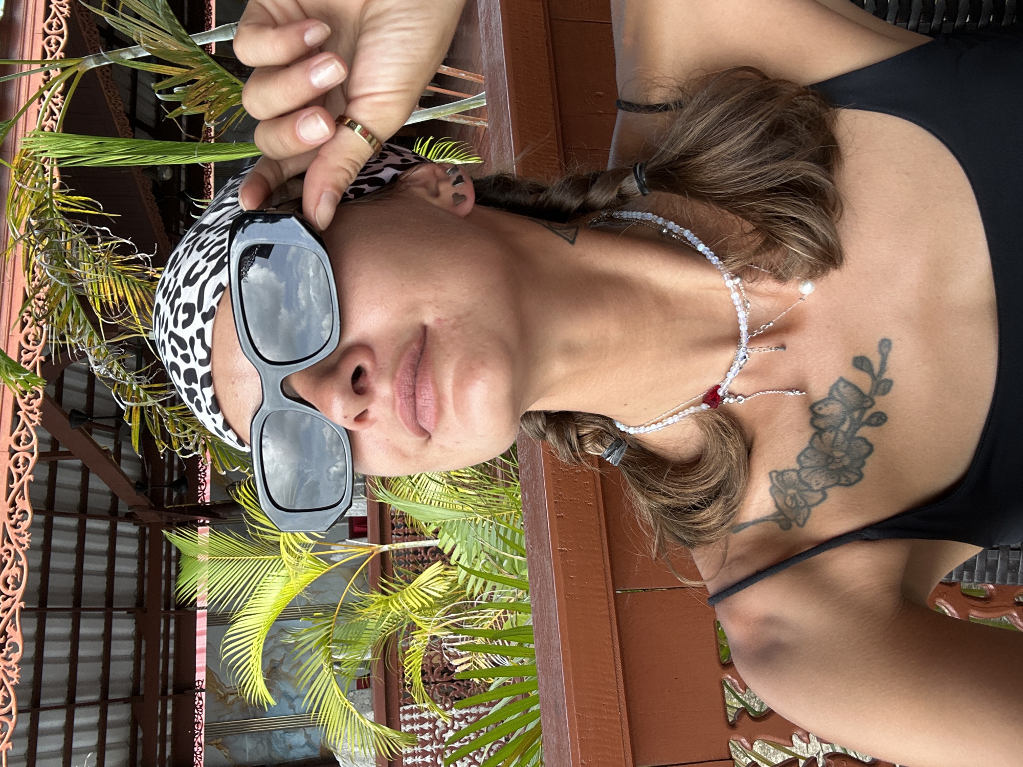 A young woman, seen from the chest up, is photographed outdoors. She is wearing a black tank top, a leopard print bandana, and large, black-framed sunglasses. She has two braids in her hair and is wearing a multi-layered necklace with beads and charms. A tattoo of flowers is visible on her left shoulder. Her right hand is raised, adjusting her sunglasses, and she is wearing a gold ring on her index finger. The background shows lush green palm trees and a wooden railing. The setting appears to be a tropical outdoor dining area with a roof structure overhead, potentially in Ko Panyi, Thailand, given the context.