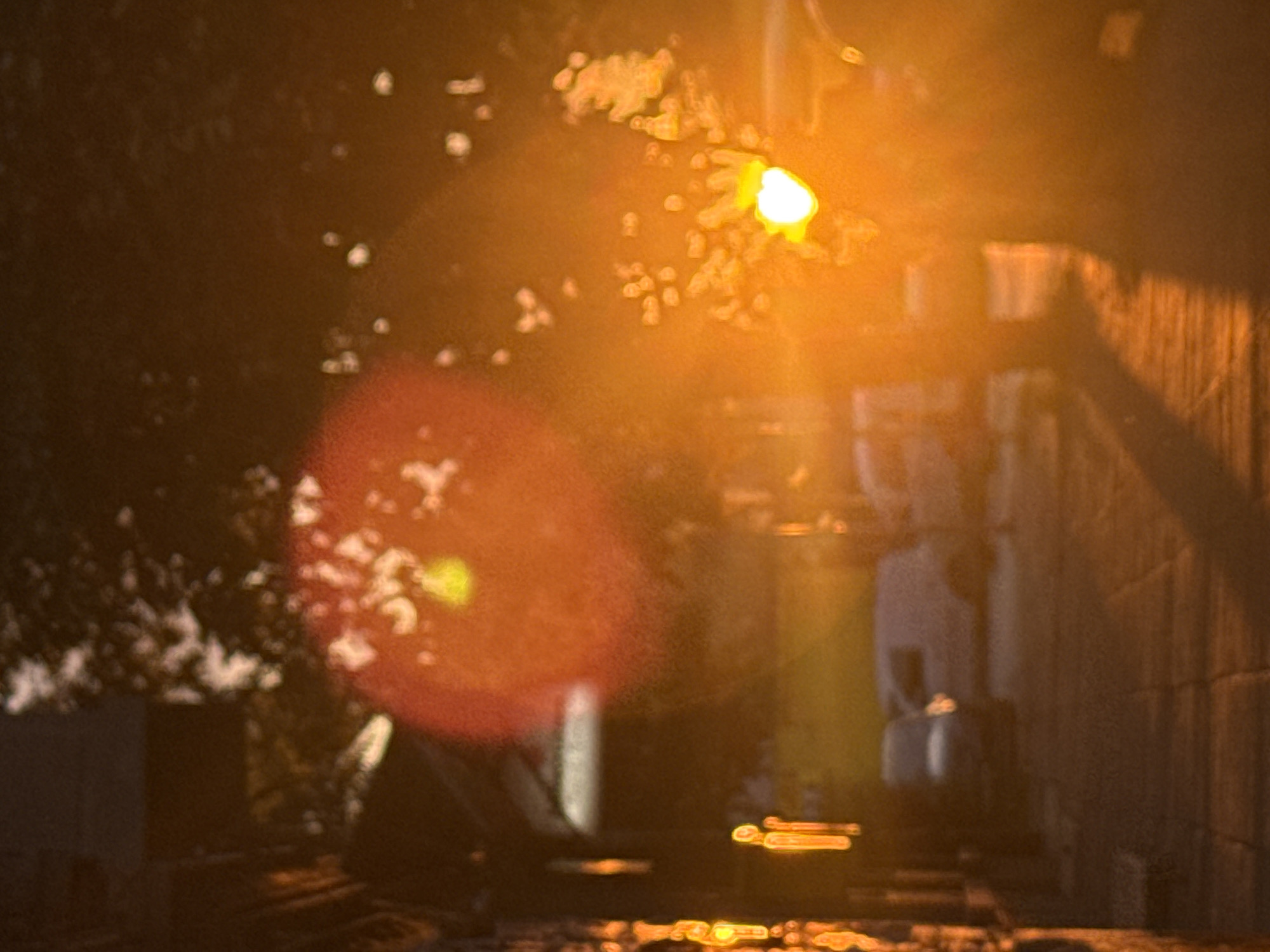 The image depicts an extremely blurred and abstract urban street scene, dominated by intense warm light and lens flares, suggestive of twilight or nighttime conditions. The predominant colors are deep orange, red, golden yellow, and dark brown.
On the far left, a vertical structure, possibly a building facade or a lamppost, shows vertical streaks of bright orange and yellow light, appearing as highly elongated, indistinct shapes. The lower-left-center shows a blurred paved surface, possibly a sidewalk or street, with subtle linear patterns.
In the mid-ground, two highly indistinct vehicles are visible. To the left-center, a darker, blurred shape suggests a sedan. Behind and slightly to its right, a lighter-colored, blurred shape is consistent with a parked or slowly moving van.
The right side of the image is characterized by an extremely bright, almost white, circular light source, causing a significant golden-orange lens flare that obscures much of the right and central portions of the frame. This light source is consistent with a strong streetlamp, a vehicle headlight, or a low sun. Above and around this intense light, dark, amorphous shapes with lighter flecks are consistent with tree foliage, backlighted or illuminated by the strong source. Another large, diffuse red-orange lens flare is prominent in the upper-mid-left section of the image.
No discernible people, specific actions, or interactions are present. The scene is too blurred to identify specific landmarks for precise geolocation within Sofia, Bulgaria.
