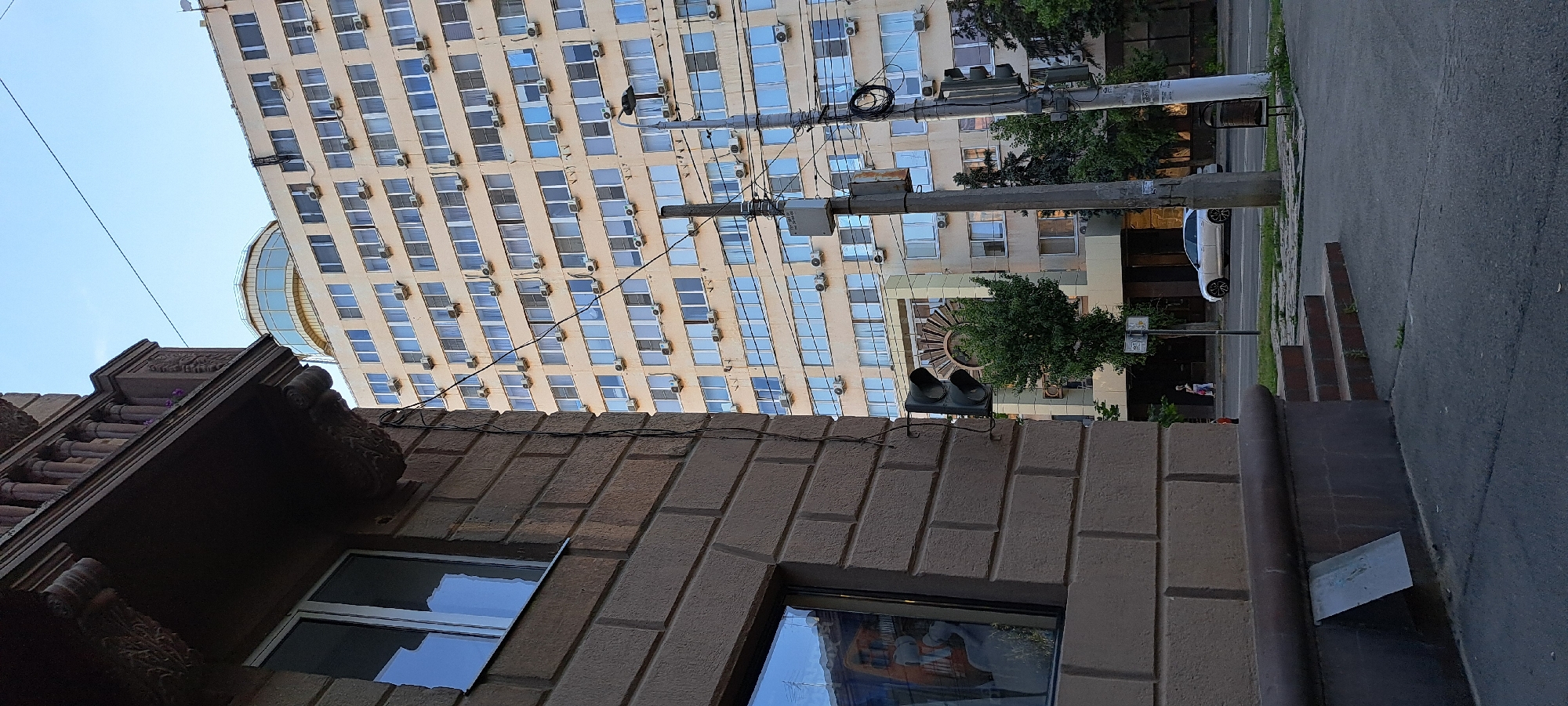 This is a medium shot, eye-level, outdoor, urban scene captured on a clear day. The foreground is dominated by the lower portion of a building with a light brown brick facade and a dark brown decorative ledge or balcony. To the left, a window on the brick building reflects the sky and some orange and grey objects. A traffic light is mounted on a pole to the right of the brick building.
In the mid-ground, a tall, light-colored apartment building with numerous windows and air conditioning units is visible. A distinctive round, glass-enclosed structure sits atop this building. A single green tree grows in front of the apartment building.
In the background, a white car is parked on the street, and a pedestrian can be seen walking. Overhead power lines and utility poles are also prominent features of the scene. The ground is paved with asphalt and concrete, with some grass growing through cracks.