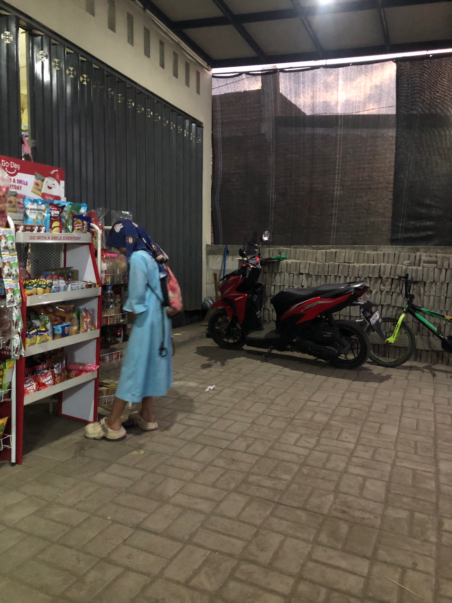 The image depicts an outdoor scene in Keniten, Indonesia. A person wearing a light blue dress and a blue hijab is standing in front of a display shelf filled with various packaged goods. The person is looking down at the shelf, and a pink and white backpack is slung over their shoulders. To the right of the person, there is a red scooter parked next to a pile of bricks. Further to the right, a green and black bicycle is leaning against the bricks. The ground is paved with interlocking rectangular bricks. In the background, a building with a dark gray corrugated metal door is visible, and above that, a dark structure with some natural light filtering through. The overall lighting suggests it is either dusk or an indoor/covered area with artificial lighting.