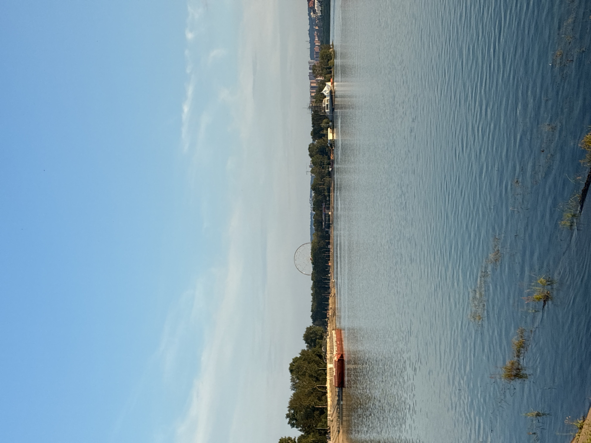 The image depicts a wide river or large body of water with a calm surface, reflecting the pale blue sky and scattered wisps of white clouds. In the foreground, shallow water shows submerged vegetation or sediment. Along the far bank, a sandy shore leads to a line of lush green trees. A red barge is moored along the bank. Further in the distance, a Ferris wheel is visible above the treeline, suggesting an amusement park or recreational area. On a small island in the middle of the water, there is a white building with an unusual, possibly futuristic, architectural design. Beyond the island, a cityscape with buildings of varying heights and colors lines the horizon. The overall scene suggests a peaceful, daytime setting, likely a waterfront park or urban recreational area in Irkutsk, Russia, near the Angara River.