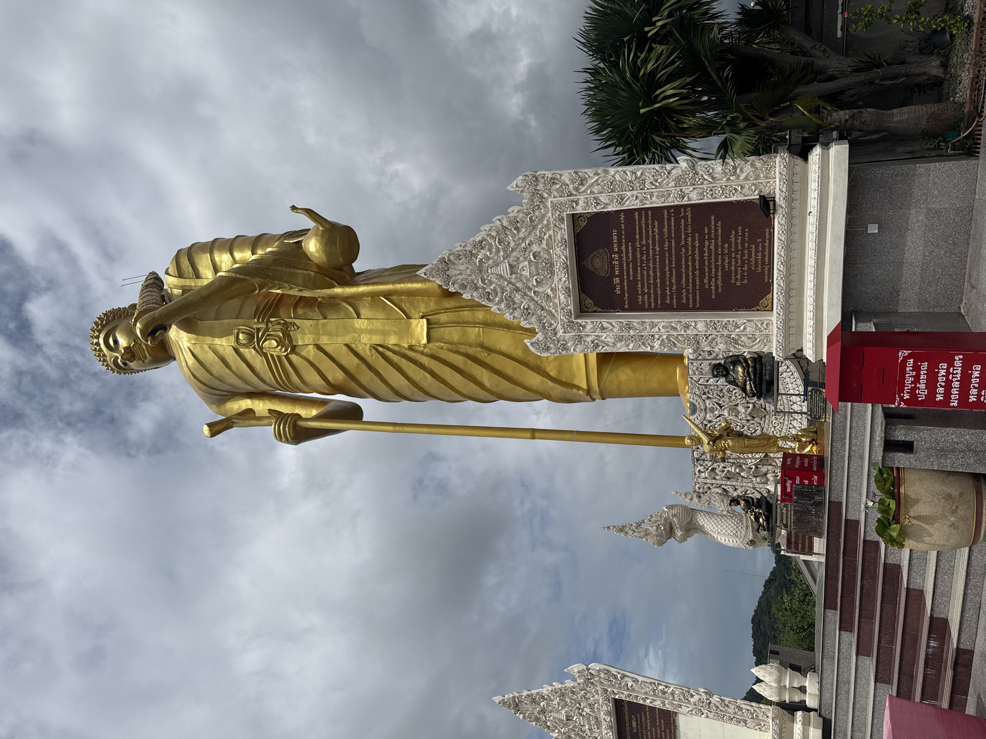 A low-angle, full-body shot captures a massive, golden Buddha statue in Hua Hin, Thailand. The statue, depicted in a standing posture, is adorned in traditional robes and holds a staff in its right hand and a vessel in its left. Its head is crowned with intricate ornamentation. The statue is situated on a raised platform with decorative white architectural elements flanking it. One of these elements contains a dark plaque with Thai script, providing information about the Buddha. In front of the platform, a red donation box is visible. The scene is set against a backdrop of a cloudy sky, with lush green trees and a hillside in the distance. The lower portion of the image shows a set of stairs leading up to the platform, with a potted plant and a glimpse of pink fabric at the bottom left.