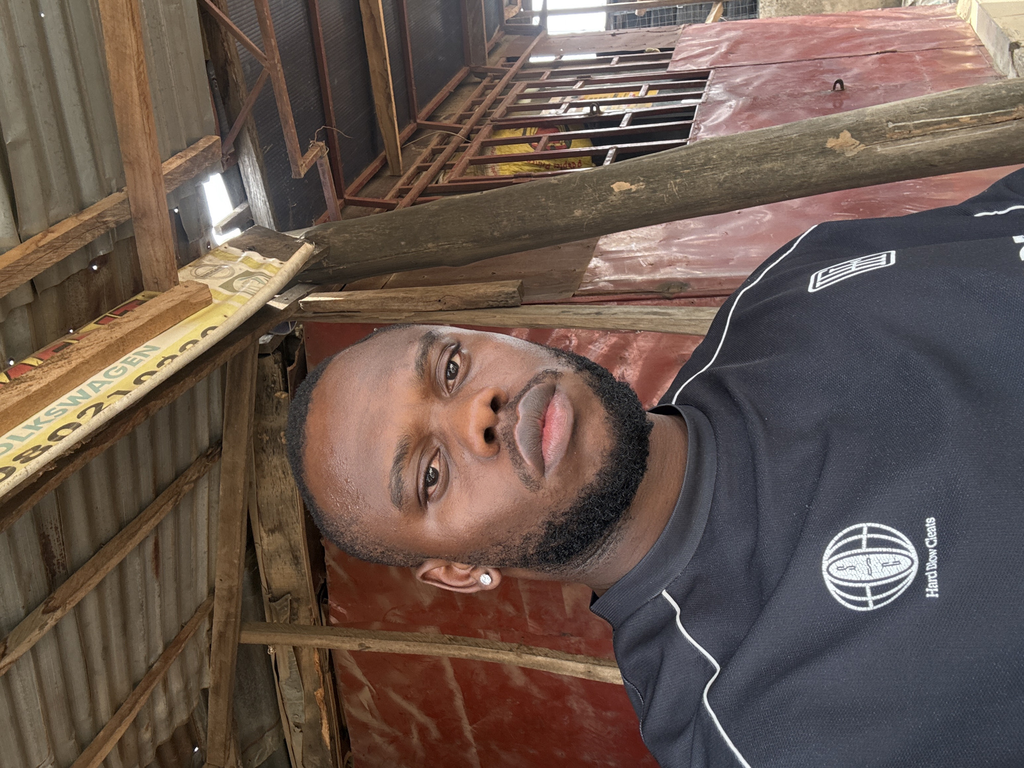 The media file presents a close-up portrait of a young Black man situated in what appears to be a rustic, partially constructed, or utilitarian indoor setting. The man has a clean-shaven head, a short, dark beard, and full lips. He looks directly at the camera with a neutral expression. A white diamond stud earring is visible in his left ear.
He is wearing a black short-sleeved top with white piping along the collar and sleeves. The shirt features a white circular logo on the right side of the chest, depicting a stylized ball (resembling a basketball or sphere with grid lines) and the text "Hard Blow Cleats" written vertically next to it.
The background consists of exposed wooden beams and planks, some corrugated metal sheets, and areas of reddish-brown painted surfaces. Bright daylight filters into the space, indicating it is daytime. On a yellow and white object in the upper left background, partially obscured by wooden beams, the word "VOLKSWAGEN" and the numbers "080210" are clearly visible, possibly part of a repurposed sign or advertisement. The setting suggests a casual environment, possibly a workshop, storage area, or a building under construction, consistent with informal structures that can be found in areas like Lugbe, Nigeria.