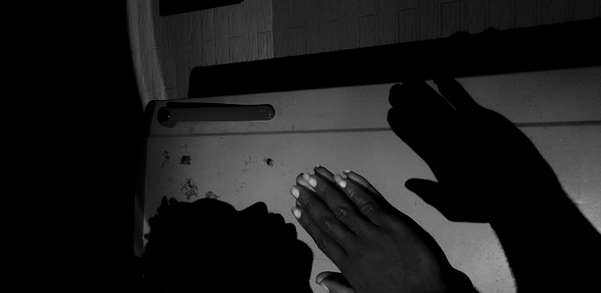 The image is a black and white photograph showing a person's hands resting on a surface, possibly a refrigerator door, in a dimly lit room. The shadow of another hand is visible beside the hands. The environment includes a wall with textured tiles behind the surface. There are marks or stains on the surface of the refrigerator door. The location of the image is Narayi, Nigeria. The subject involves the interaction of a person with an object, likely in a domestic setting.