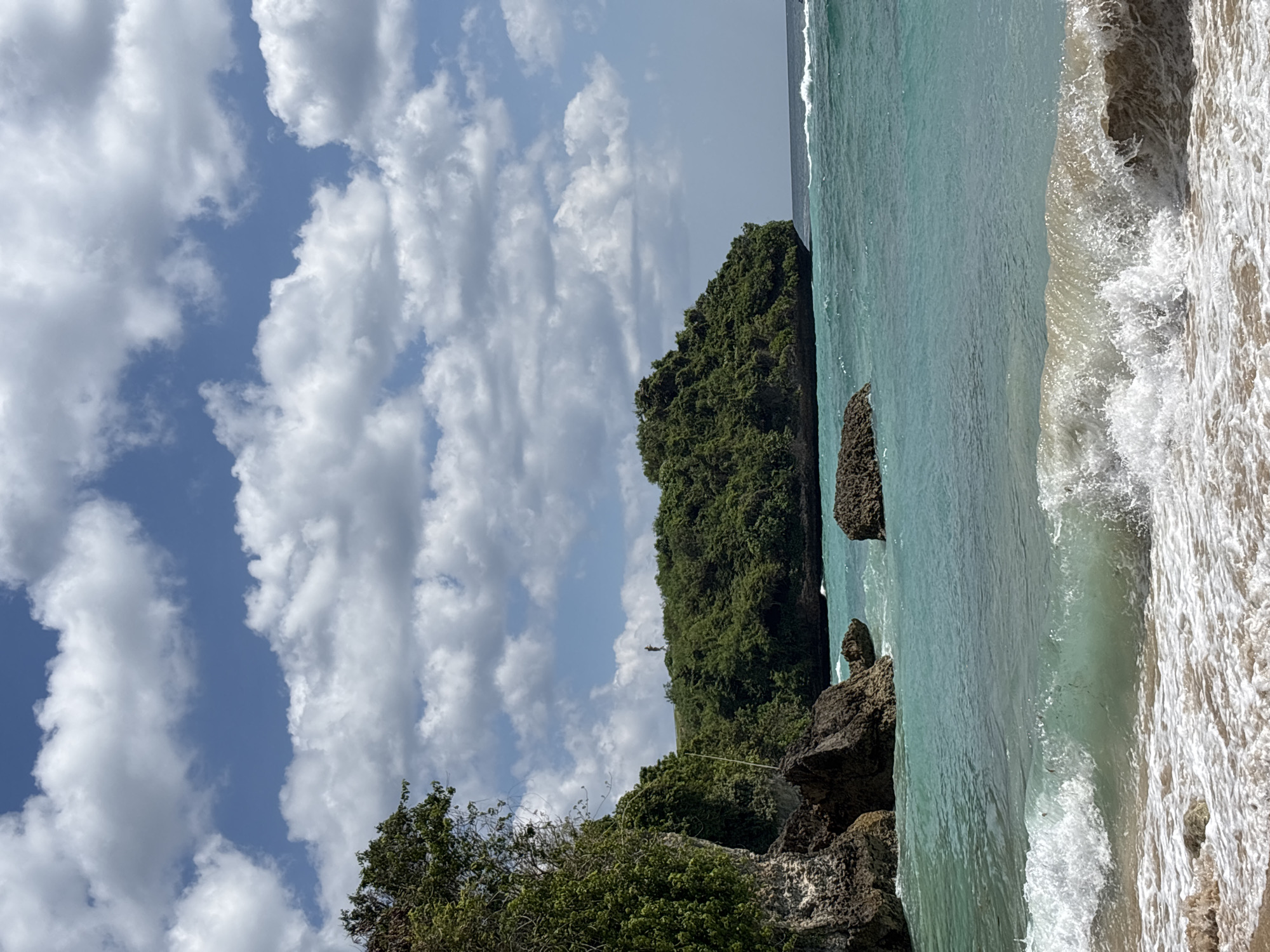 This image displays a coastal landscape in Kuta Selatan, Indonesia. The foreground features a sandy beach with white-capped waves breaking and receding, creating foam on the light brown sand. The ocean transitions from a vibrant turquoise near the shore to a deeper blue further out, with additional wave activity visible towards the horizon. To the left, a steep, rocky cliff face is densely covered with lush green trees and various vegetation. Multiple dark rock formations are present along the shoreline and within the shallow water. In the mid-ground, a significant, rounded landmass or island, entirely covered in dense green foliage, extends into the ocean. A solitary human figure, observed as a distant silhouette, stands upright on the elevated crest of this verdant headland. The sky above is clear blue with numerous scattered, fluffy white cumulus clouds.