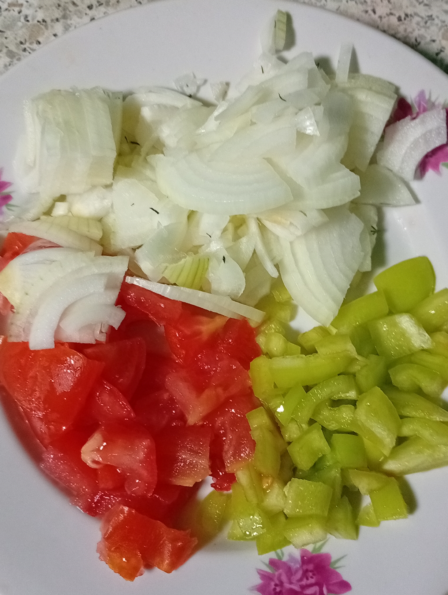 A white plate contains chopped ingredients: sliced white onions, diced red tomatoes, and diced green bell peppers. The plate is adorned with floral patterns. The background appears to be a speckled countertop. The image is an overhead shot.