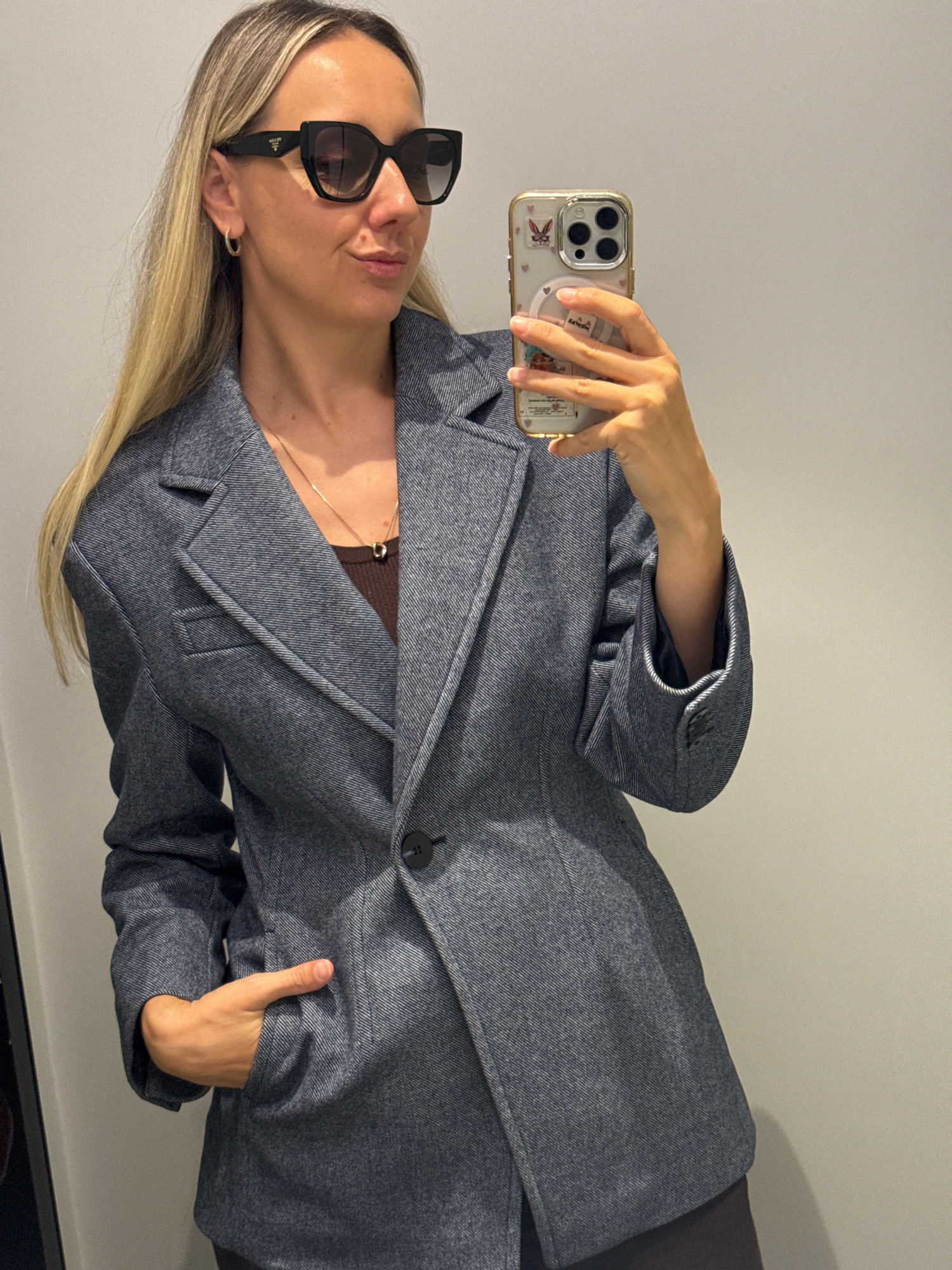 A blonde woman with large black sunglasses and a gold necklace stands in what appears to be a fitting room, holding a smartphone and taking a selfie. She is wearing a dark grey blazer over a brown top. The background is a plain, light-colored wall, suggesting an indoor environment like a retail store or a home. There are no visible landmarks or clues to indicate a specific location in Riga, Latvia.