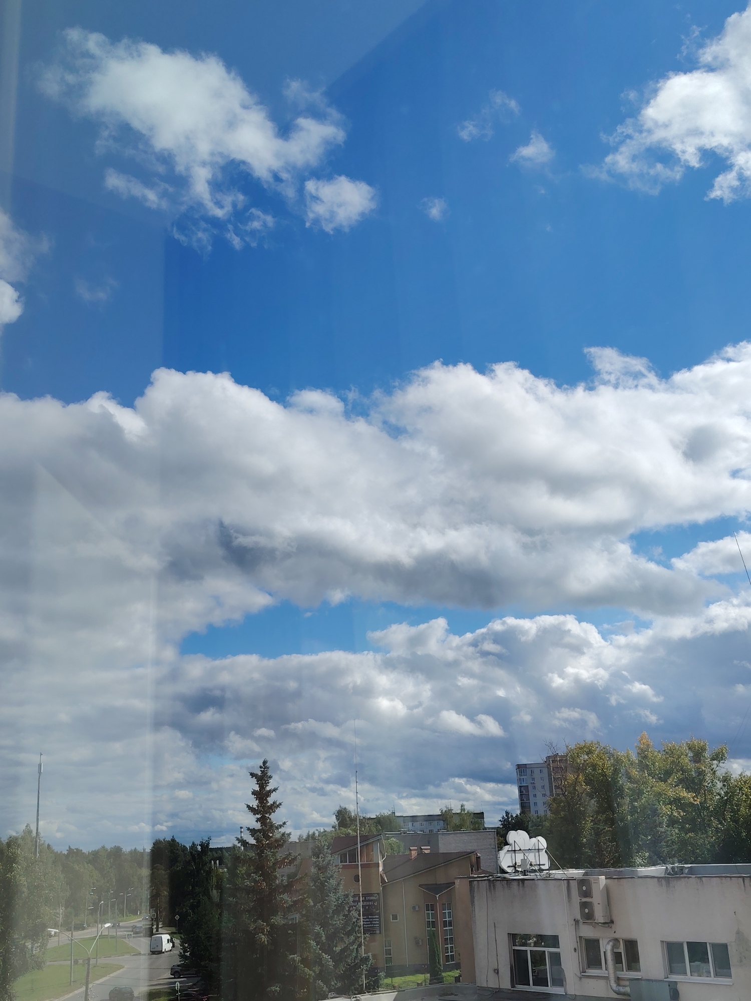 The image shows a view from a window looking out onto a city in Konakovo, Russia, on a partly cloudy day. The sky is filled with large, puffy white clouds against a bright blue backdrop. Below the sky, a dense line of green trees forms the horizon. In the foreground, there are several buildings of varying heights and architectural styles. To the left, a road with parked cars and a white van is visible. A tall, slender evergreen tree stands prominently in the center of the frame, with a few other trees and buildings scattered around it. Some of the buildings have satellite dishes and air conditioning units on their roofs. The overall scene depicts a typical urban landscape with elements of nature.