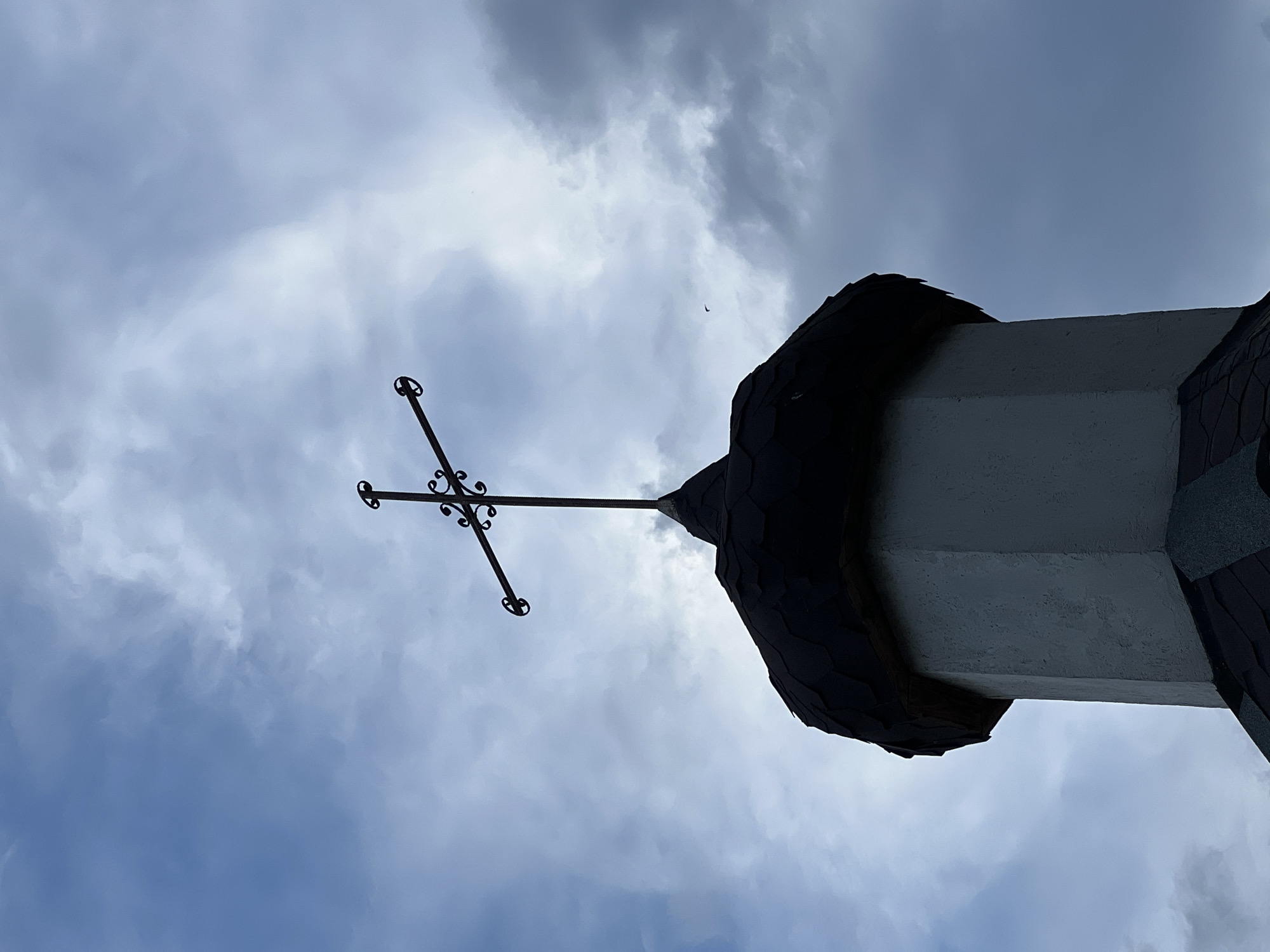 An ornate wrought-iron cross tops a hexagonal steeple covered in dark shingles. The steeple, a light gray color, is part of an octagonal structure. The sky above is overcast with dramatic, moody clouds, suggesting an approaching storm or a transition in weather. The overall scene is stark and silhouetted, with the cross and steeple sharply defined against the turbulent sky. The image is taken from a low angle, emphasizing the height of the structure and the vastness of the sky. There are no people visible in the image. This structure is located in Năvodari, Romania.