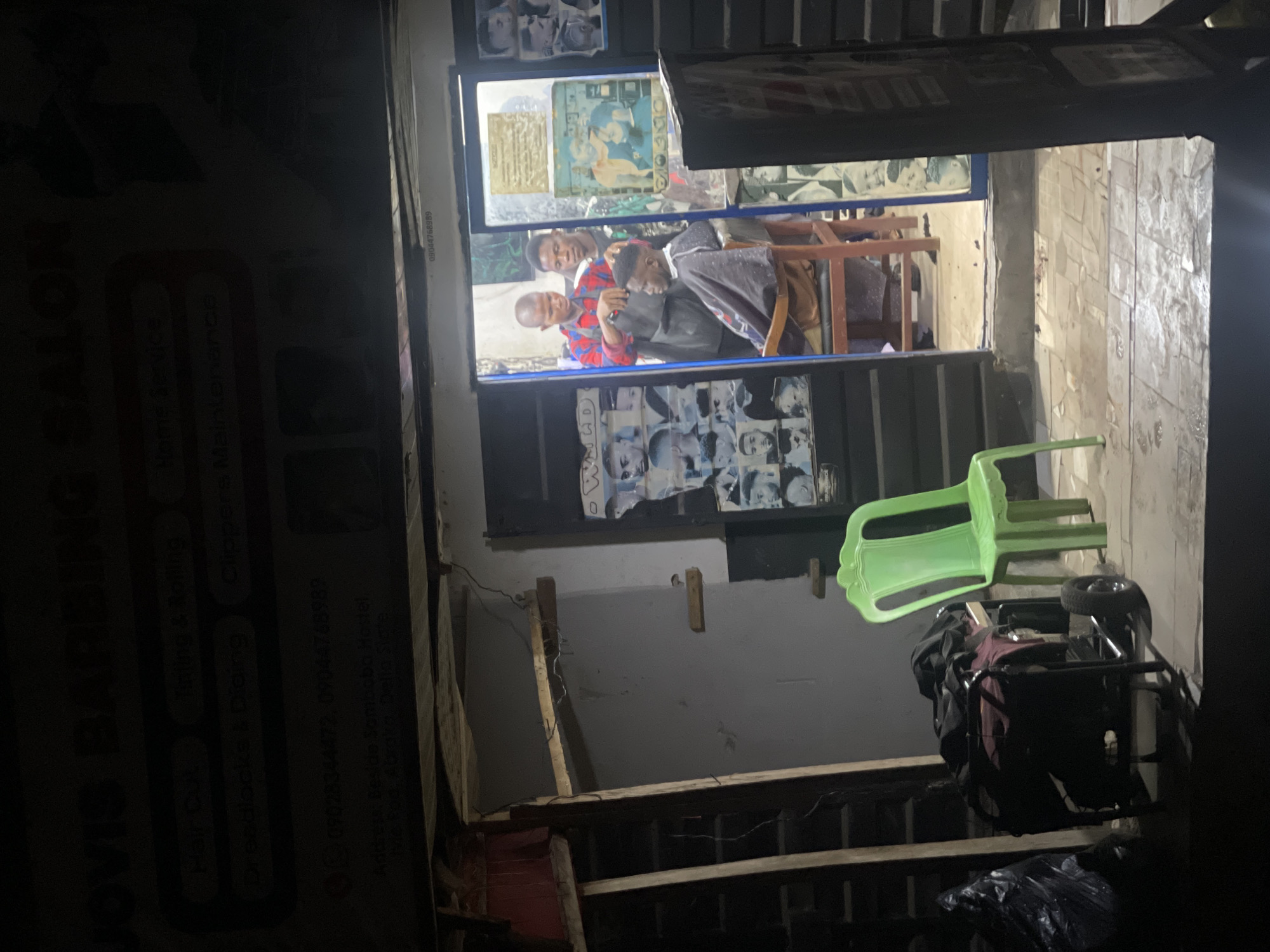 The image shows a dimly lit barbershop in what appears to be a low-income area, likely in Abraka, Nigeria. Through the doorway, a barber is cutting a customer's hair. Two other people are present in the shop. Outside the shop, there is a green plastic chair and a pushcart. The shop's sign, partially visible on the left, advertises "Dovis Barbering Salon," offering services such as "Hair Cut," "Tinjing & Rolling," "Home Service," "Clippers Maintenance," and "Dreadlocks & Diding." Contact phone numbers (09028344472, 09044768989) are listed. The address is "Beside Sambobn Hostel." The surrounding environment appears somewhat dilapidated, with unfinished construction materials visible in the foreground.
