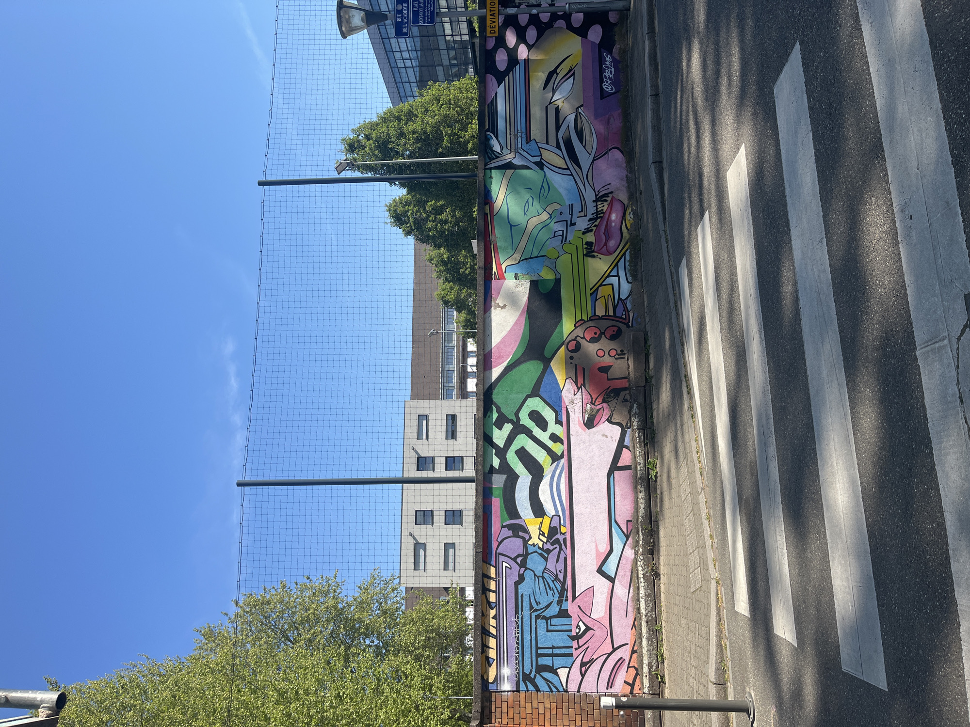 The image shows a section of street art painted on a retaining wall along a sidewalk in Strasbourg, France. The style is colorful and cartoonish, with various figures and abstract shapes. The sidewalk is adjacent to a road with a zebra crossing. A metal fence runs along the top of the wall. In the background, buildings and trees are visible. The sky is clear and blue. Based on the street sign visible in the image, the location is near Rue de l'Académie in Strasbourg.