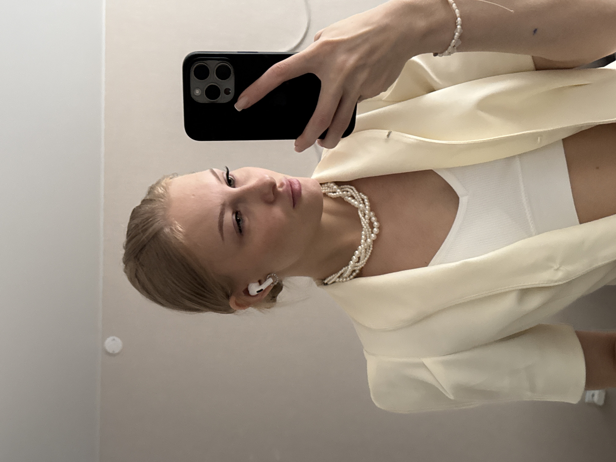 A young woman with blonde hair pulled back is taking a mirror selfie indoors. She is wearing a white blazer over a white tank top and a multi-strand pearl necklace. She has white wireless earbuds in her ears and a pearl bracelet on her right wrist. She is holding a black smartphone in her right hand, capturing her reflection. The background appears to be a wall with a subtle texture and a portion of a ceiling is visible at the top. There is no indication of the city of Perm, Russia, or any specific urban environment within the image.