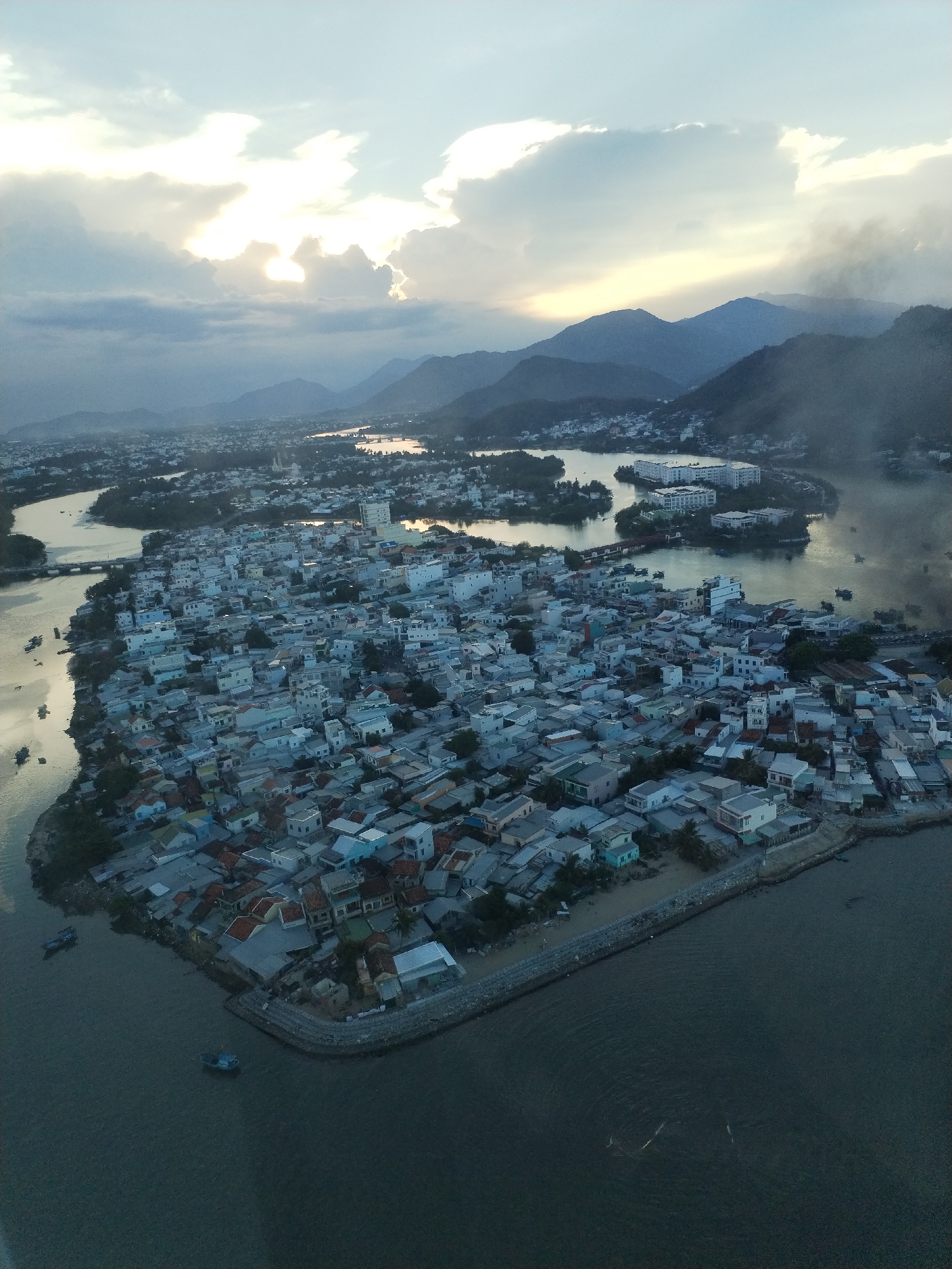 The image shows an aerial view of Bac Nha Trang Ward, Vietnam. The scene is dominated by a dense collection of low-rise buildings, primarily residential, covering a peninsula extending into a river or bay. Many buildings have tiled roofs, some with faded or colorful hues. The peninsula is bordered by a stone seawall. The water reflects the sunlight. In the background, mountains are visible. The sky is overcast with clouds, and sunlight is filtering through in the upper part of the image. Boats are scattered in the waters around the peninsula.