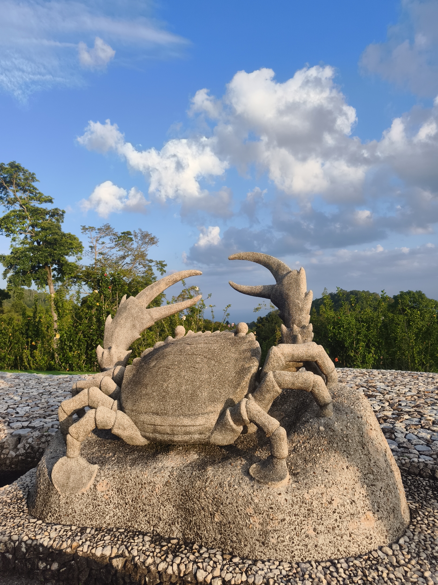 A stone sculpture of a crab rests on a large boulder, surrounded by a ground covered in small stones. The crab sculpture is intricately detailed, with visible legs, claws, and body segments. Behind the sculpture, lush green trees and vegetation form a dense backdrop, indicating a natural or park-like setting. In the distance, a hazy horizon suggests a broader landscape, possibly with hills or mountains. The sky above is a clear blue, punctuated by scattered white clouds, illuminated by what appears to be sunlight. This scene is located in the Koh Samui District of Thailand.