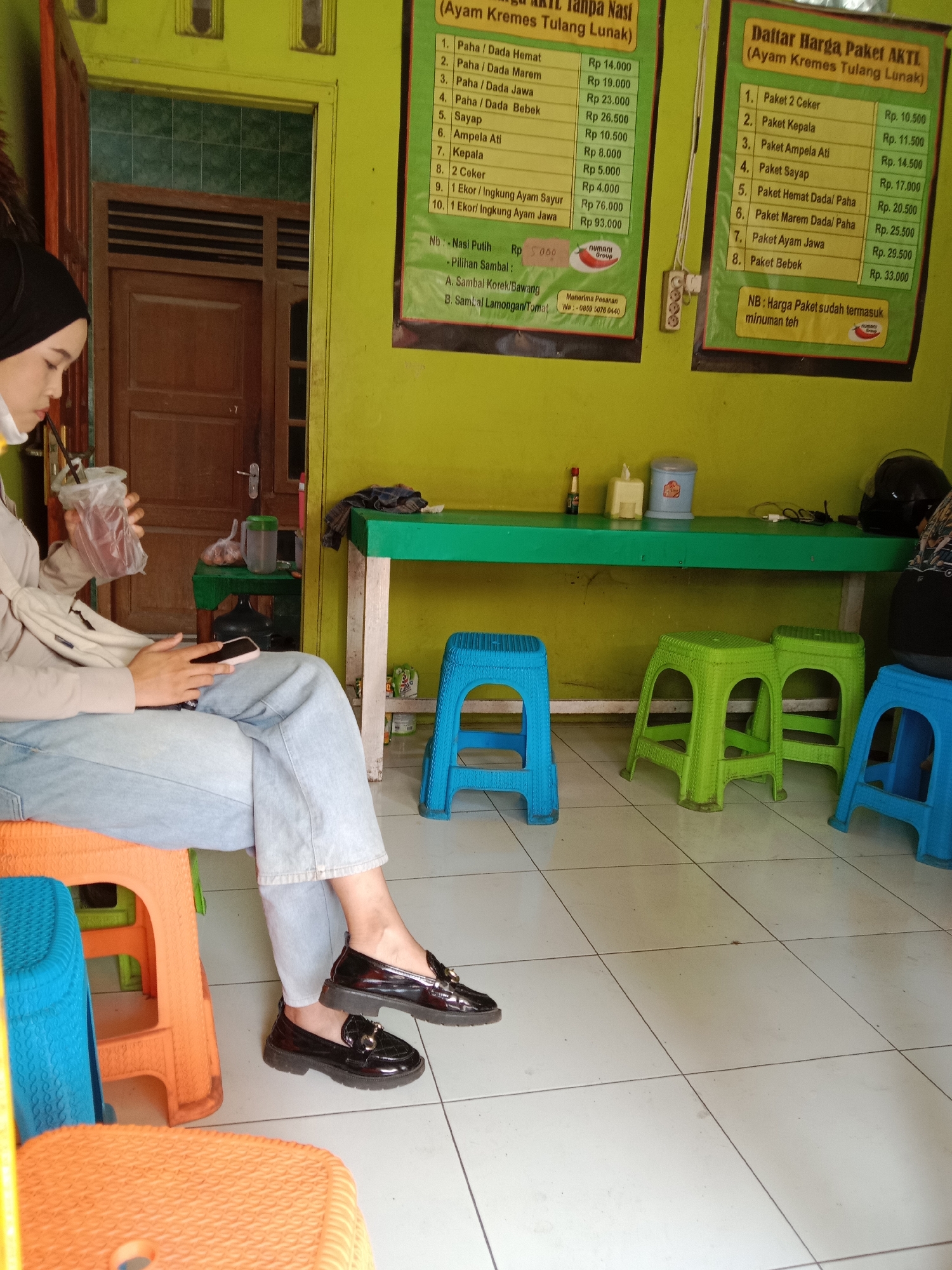 The image shows a woman seated indoors at what appears to be a casual eatery or waiting area, characterized by bright lime-green walls and a white tiled floor. The woman, positioned on the left, wears a black hijab, a white face mask pulled below her chin, a light-colored long-sleeved top, light blue wide-leg jeans, and black platform loafers with gold accents. She holds a transparent plastic cup with a dark beverage and a straw in her right hand, while her left hand is occupied with a pink smartphone.
Various plastic stools in orange, blue, and green are scattered on the floor. A long green table against the wall holds items such as a red condiment bottle, a yellow tissue dispenser, a light blue container, a black motorcycle helmet, and a dark cloth. A smaller green table near a wooden door and window in the background holds a clear bottle and a plastic bag.
Two price lists, written in Indonesian, are displayed on the green wall. The left menu, titled "Tanpa Nasi (Ayam Kremes Tulang Lunak)", lists individual items and prices (e.g., "Paha/Dada Hemat" for Rp 14.000, "Sayap" for Rp 10.500, "1 Ekor/Ingkung Ayam Jawa" for Rp 93.000), "Nasi Putih Rp 5000", "Pilihan Sambal", and a WhatsApp number (0859 5076 0440) for orders, featuring a "NUMANI Group" logo. The right menu, "Daftar Harga Paket AKTL (Ayam Kremes Tulang Lunak)", details package deals (e.g., "Paket 2 Ceker" for Rp 10.500, "Paket Kepala" for Rp 11.500, "Paket Bebek" for Rp 33.000) and notes that package prices include tea. The scene takes place during the daytime, and the woman appears engrossed in her phone, likely waiting or taking a break.