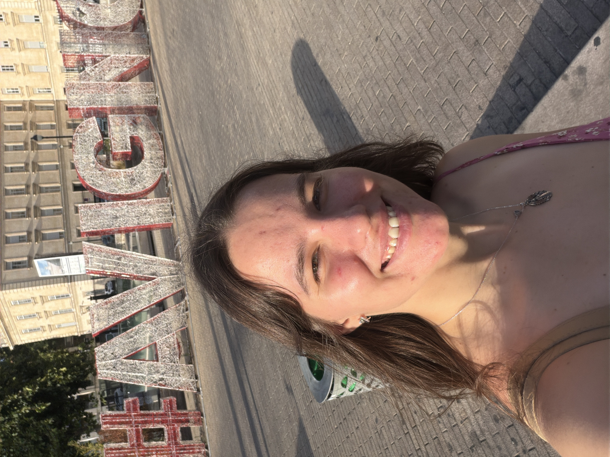 In the city of Avignon, France, a young woman with long brown hair takes a selfie in front of a large, illuminated sign that reads "#AVIGNON." The sign is made of metal letters outlined with white lights and a red backing, standing tall on a paved area. Behind the sign, a multi-story building with light-colored walls and numerous windows is visible, casting a shadow on the ground. To the left of the woman, a trash can with a green interior and a metallic exterior is situated. The woman, wearing a floral print dress and a silver necklace with a pendant, smiles at the camera. The sunlight casts a long shadow on the ground, indicating it is late afternoon or early evening.