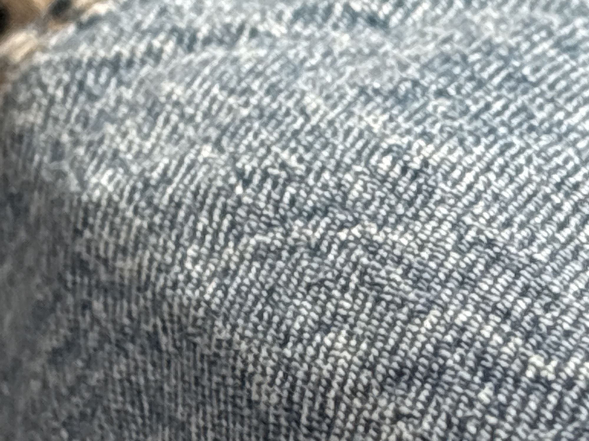This is a close-up, macro photograph of denim fabric. The image is tightly cropped, focusing solely on the texture and weave of the material. The denim appears to be a classic blue, with variations in shade due to the light catching the raised threads of the twill weave. The diagonal lines of the weave are clearly visible, creating a distinct herringbone pattern. Some areas of the fabric appear slightly creased, indicating it might be part of clothing or upholstery. The lighting is even, highlighting the texture without harsh shadows. There are no people, objects, or discernible setting other than the fabric itself. The image is static, with no activity taking place. There is no visible text in the image.