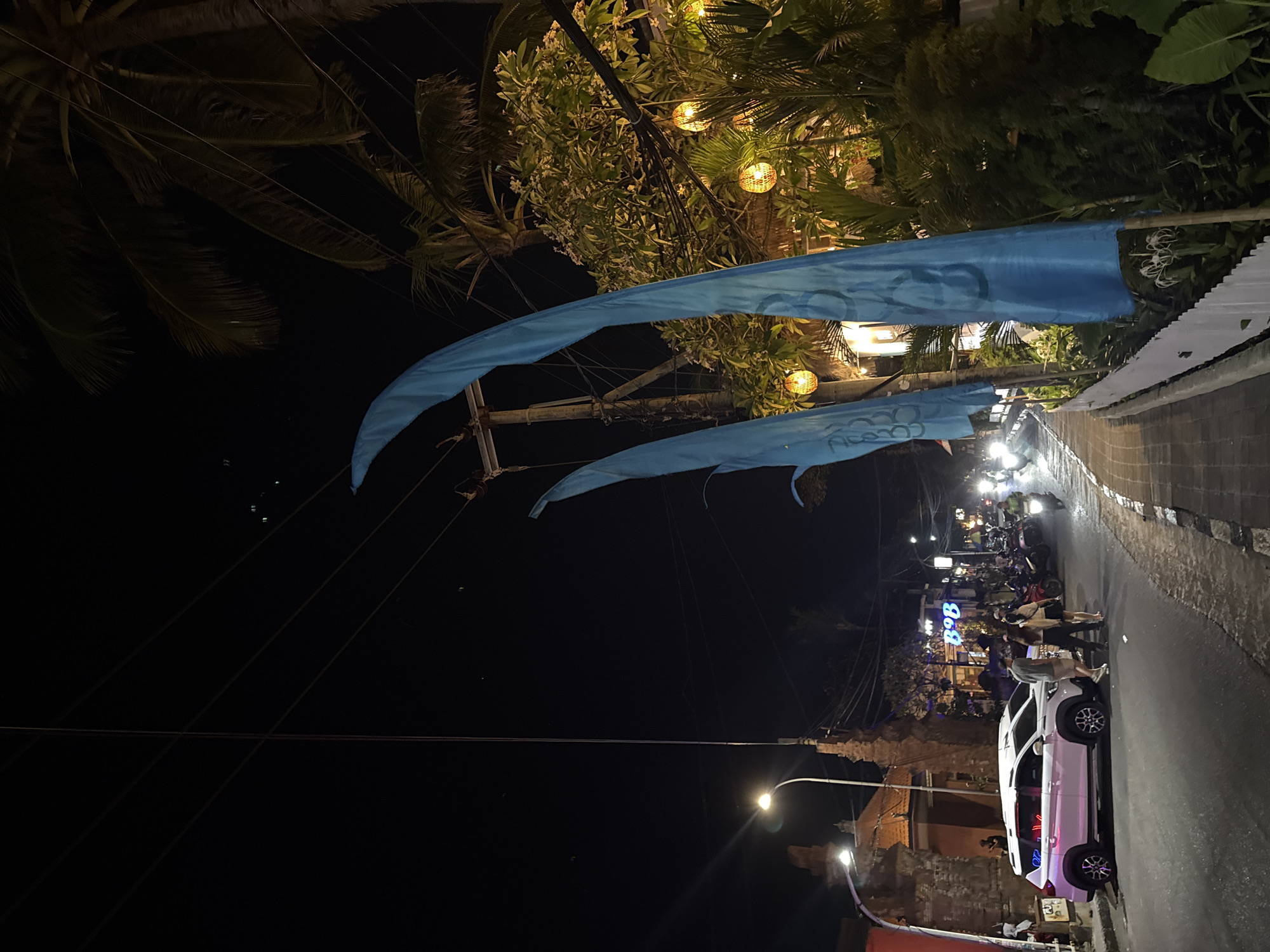 The image depicts a nighttime street scene in Kuta, Indonesia. A paved road with a sidewalk runs diagonally from the lower left to the upper right of the frame. On the left side of the road, a white SUV with pink neon lights on its side is parked. Several people are standing near the SUV, and a few motorcycles are parked further down the street. On the right side of the sidewalk, large, bright blue flags are prominently displayed. Behind the flags, lush greenery is visible, illuminated by several hanging lanterns with a woven texture, suggesting a restaurant or cafe. Palm trees are present in the background, with power lines crisscrossing the dark sky. The overall atmosphere is that of a lively street, likely a tourist area, at night.