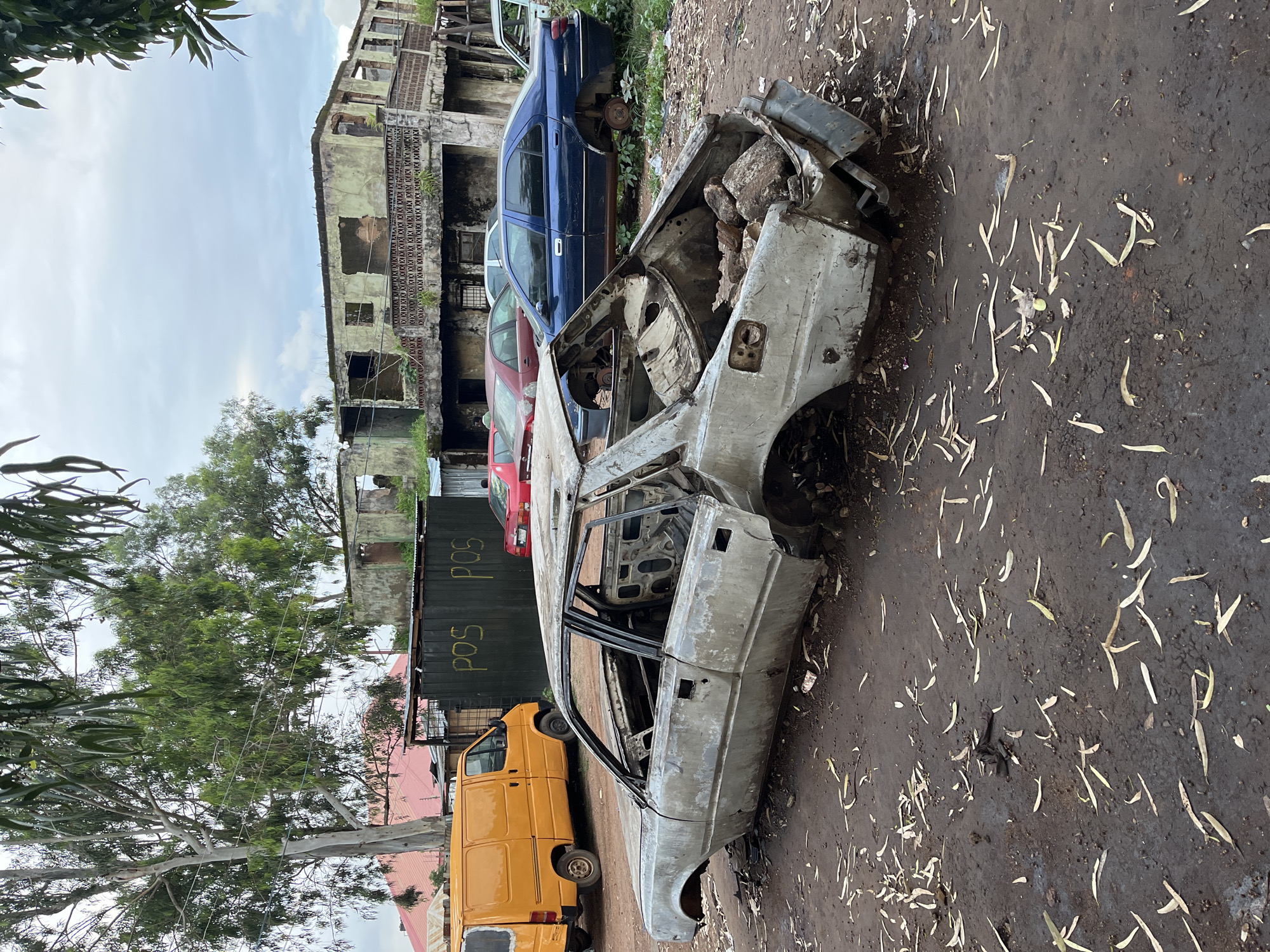 In Bukuru, Nigeria, a derelict silver sedan lies on a muddy ground covered with fallen leaves. The car is stripped of its doors, hood, and trunk, with its interior exposed and filled with debris and large rocks. In the background, several other vehicles are parked, including a yellow van, a red car, and a blue sedan. Behind the vehicles, a dilapidated two-story building with broken windows and crumbling walls stands under a cloudy sky. The scene appears to be an open lot or junkyard, characterized by the presence of abandoned vehicles and decaying structures.