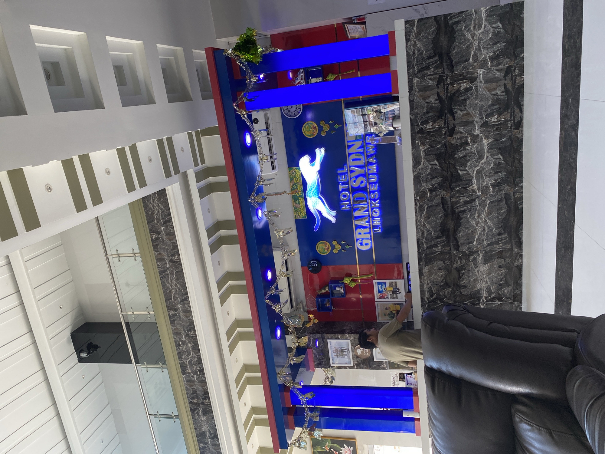 A man stands behind a reception desk at the Hotel Grand Sydn in Lhokseumawe, Indonesia. The hotel lobby features a prominent blue and red reception counter decorated with silver streamers. A large sign behind the counter reads "HOTEL GRAND SYDN LHOKSEUMAWE" and features a stylized white jaguar. The man is wearing a tan t-shirt and appears to be interacting with the desk. The lobby is well-lit with recessed ceiling lights and accent lighting on the reception counter. A black leather armchair is visible in the foreground. The walls have a dark marble-like pattern.