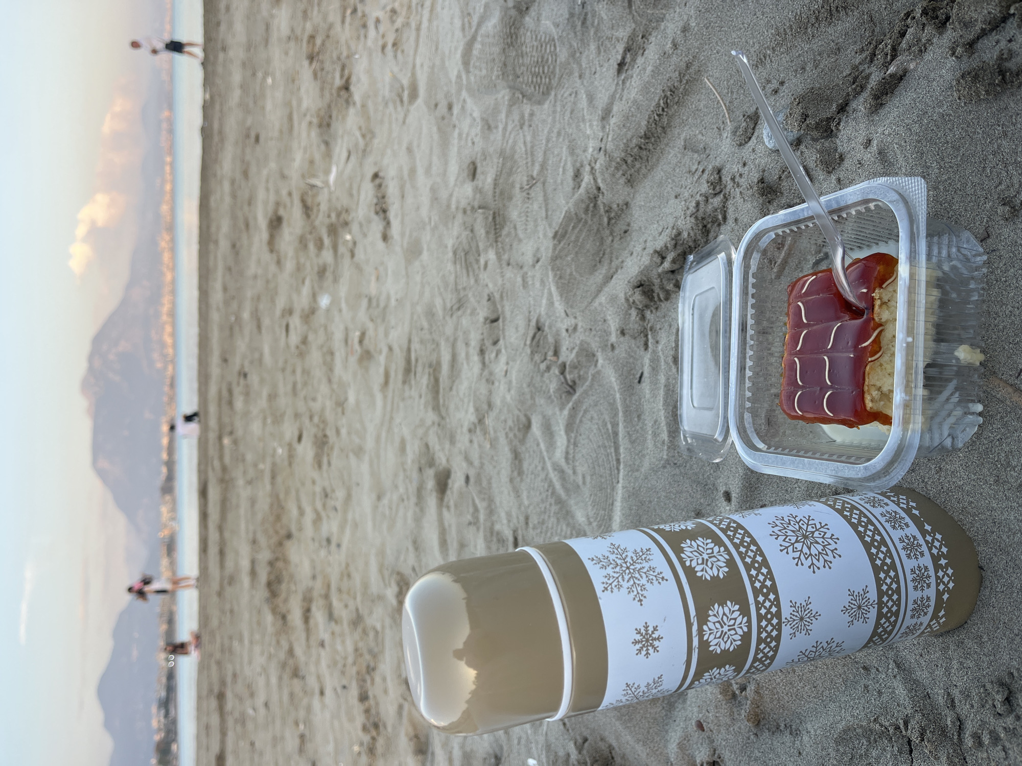 The image captures a peaceful beach scene, likely during the late afternoon or early evening as indicated by the soft light and distant glows. In the foreground, resting on light brown sand, are two main subjects: a dessert and a thermos. The dessert is a slice of what appears to be a cake or pastry, visible in a clear plastic container with an open lid. It features a reddish-brown glossy glaze with decorative white lines, and a transparent plastic fork is inserted into it. Beside the dessert is a cylindrical thermos, colored beige with a prominent white band adorned with a repeating pattern of white snowflakes and geometric designs. The sand shows various textures and faint footprints.
In the background, a wide expanse of water, presumably the sea, stretches towards a distant coastline. Beyond the water, large, dark mountains rise, meeting a light sky. Several blurred figures of people are visible along the distant shoreline, scattered across the beach or near the water's edge. A subtle golden glow, possibly from early city lights, can be seen at the base of the mountains along the coast. The scene suggests a relaxing moment, perhaps a snack or picnic, by the sea. No visible text is present on any of the objects in the image. The setting aligns with coastal locations such as Alanya, Turkey, which features mountains meeting the sea.