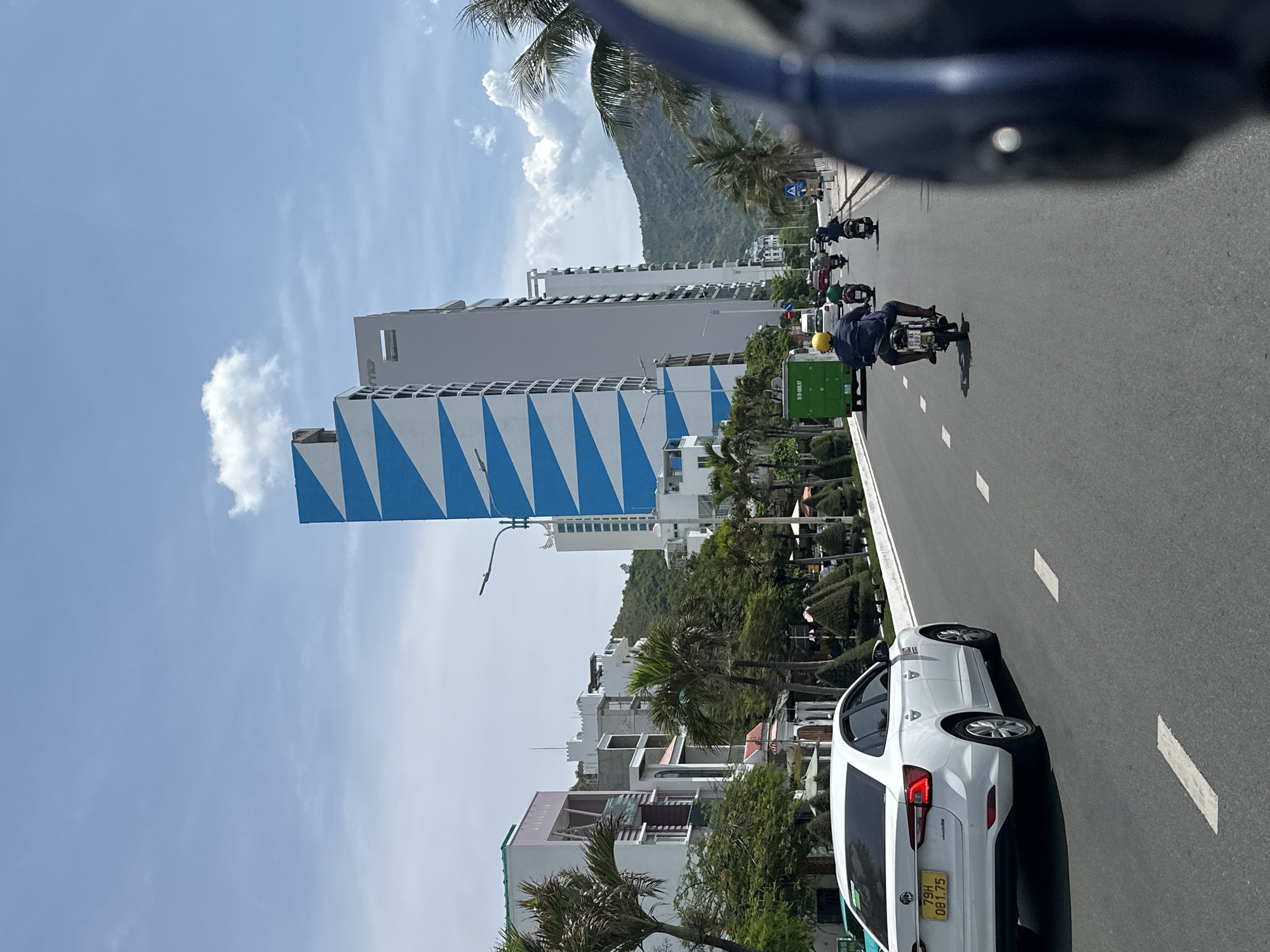 The image depicts a daytime scene on a street in Nha Trang, Vietnam, with a white car in the foreground, its license plate reading "79H 081.75." Several motorcycles are in motion on the road, with one rider wearing a yellow helmet. Tall buildings line the street, including a prominent white building with a distinctive blue zigzag pattern. Greenery and palm trees are visible, and a lush green mountainside forms the backdrop. The sky is partly cloudy. The perspective suggests the image was taken from a moving vehicle, possibly a motorcycle, given the view of a helmet in the lower right corner.