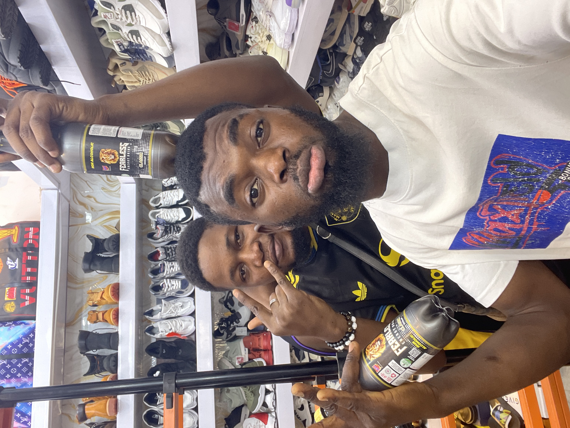 Two young men pose in what appears to be a shoe store. One wears a white t-shirt with a blue design; the other wears a black Adidas t-shirt with yellow stripes. Both hold bottles of "Fearless" energy drink. Shelves of shoes line the background, including sneakers, boots, and sandals. A Louis Vuitton bag is visible on a shelf. This photo may have been taken in Jos, Nigeria, due to the presence of a bottle of Fearless Energy Drink.