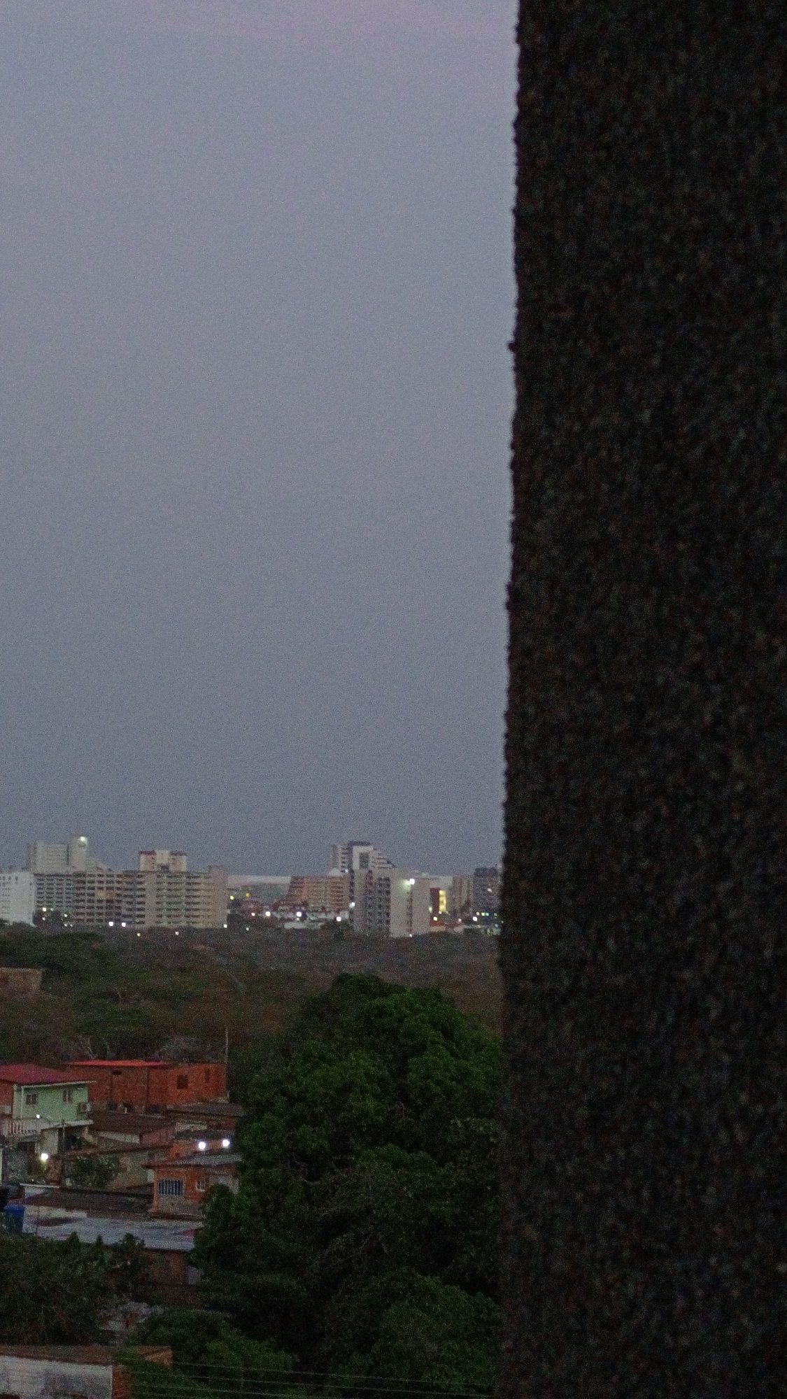 The image shows an urban landscape at dusk, likely in Los Robles, Venezuela. In the foreground, residential buildings and greenery are visible, with some structures having lights on. In the background, there is a skyline of taller buildings, also lit, suggesting a denser urban area. A dark, textured vertical element occupies the right side of the frame, partially obscuring the view. The sky is overcast and grey.
