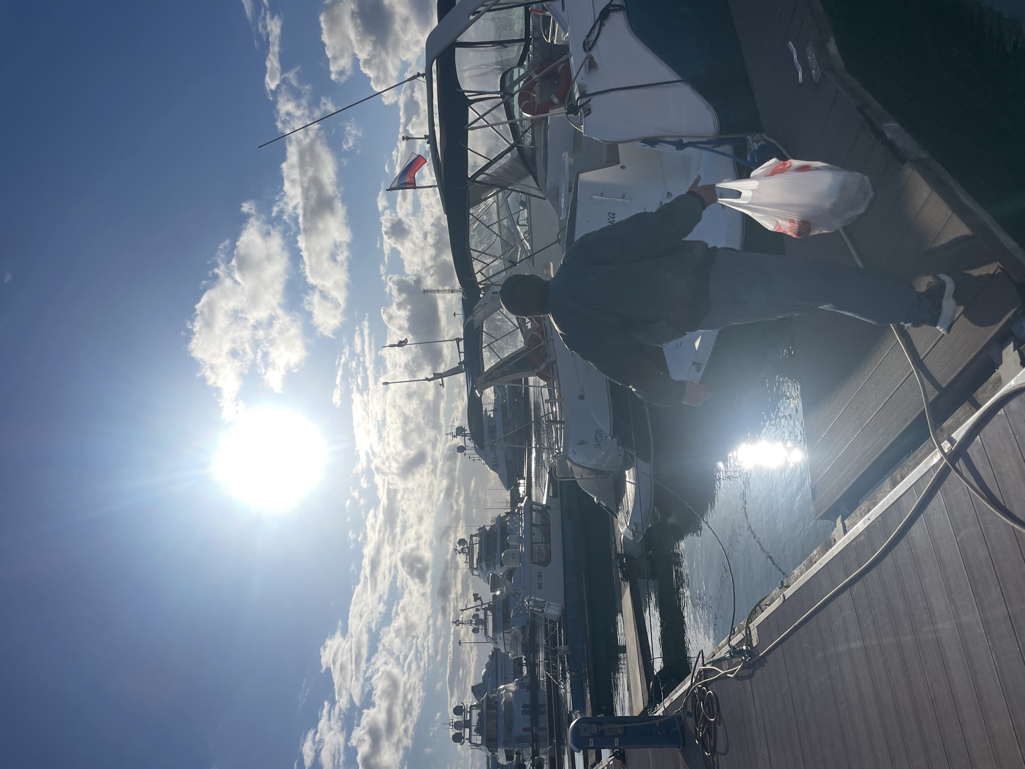 A man stands on a wooden dock, holding a white plastic bag, near a boat in the city of Sochi, Russia. Multiple boats are docked in the water. A Russian flag is seen on one of the boats. The sky is blue with white clouds, and the sun is shining brightly.