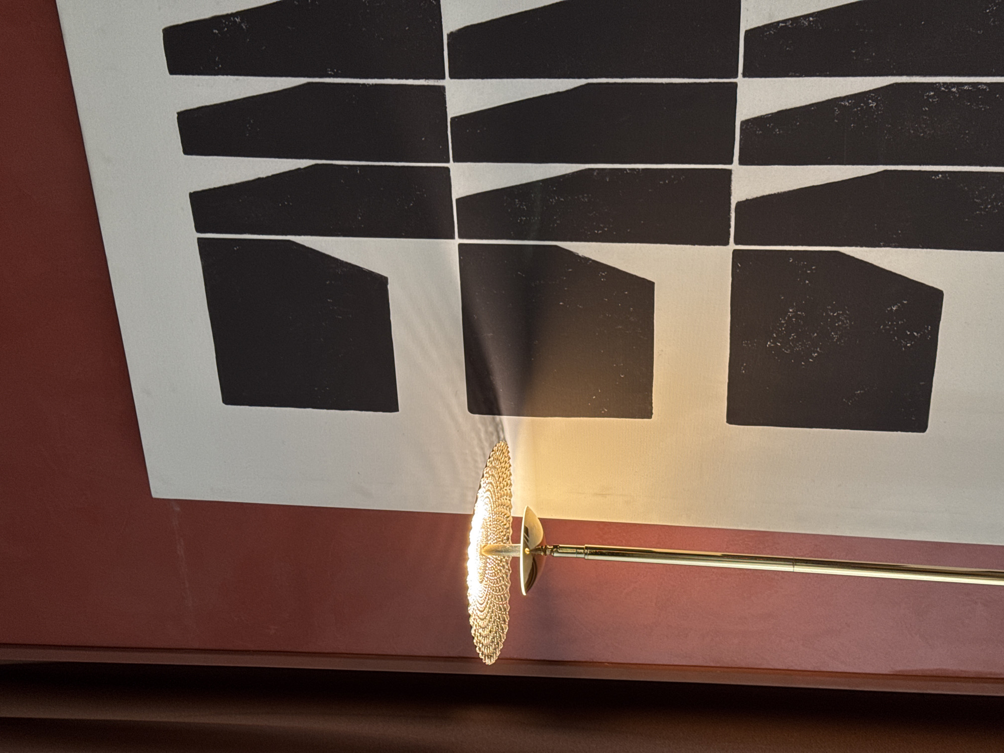 A close-up indoor shot depicts a wall lamp illuminating a framed piece of art. The artwork features a pattern of black geometric shapes against a white background, set within a red-brown frame. The lamp has a long, slender brass arm extending from a round, decorative shade that emits a bright light. The image provides no information to determine the location is Yerevan, Armenia.