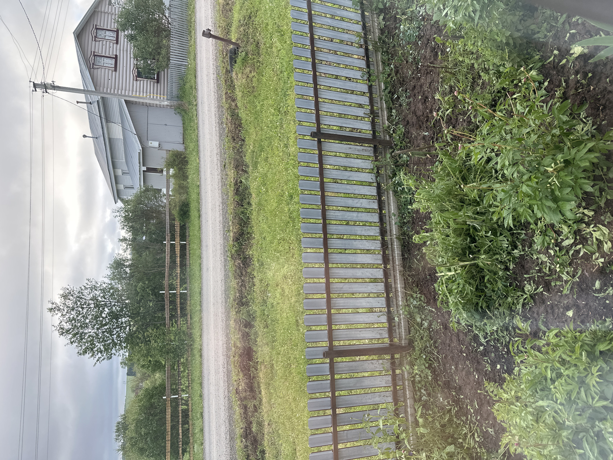 The image depicts a rural scene in Среднее Кечево, Russia. A gravel road runs parallel to a wooden fence on the left side of the frame. Beyond the road is a grassy area with sparse vegetation, a wooden fence, trees, and power lines. On the left is a wooden house with decorative window frames. To the right of the wooden fence is a cultivated garden bed with plants growing. The sky is overcast. There are no people visible in the image.