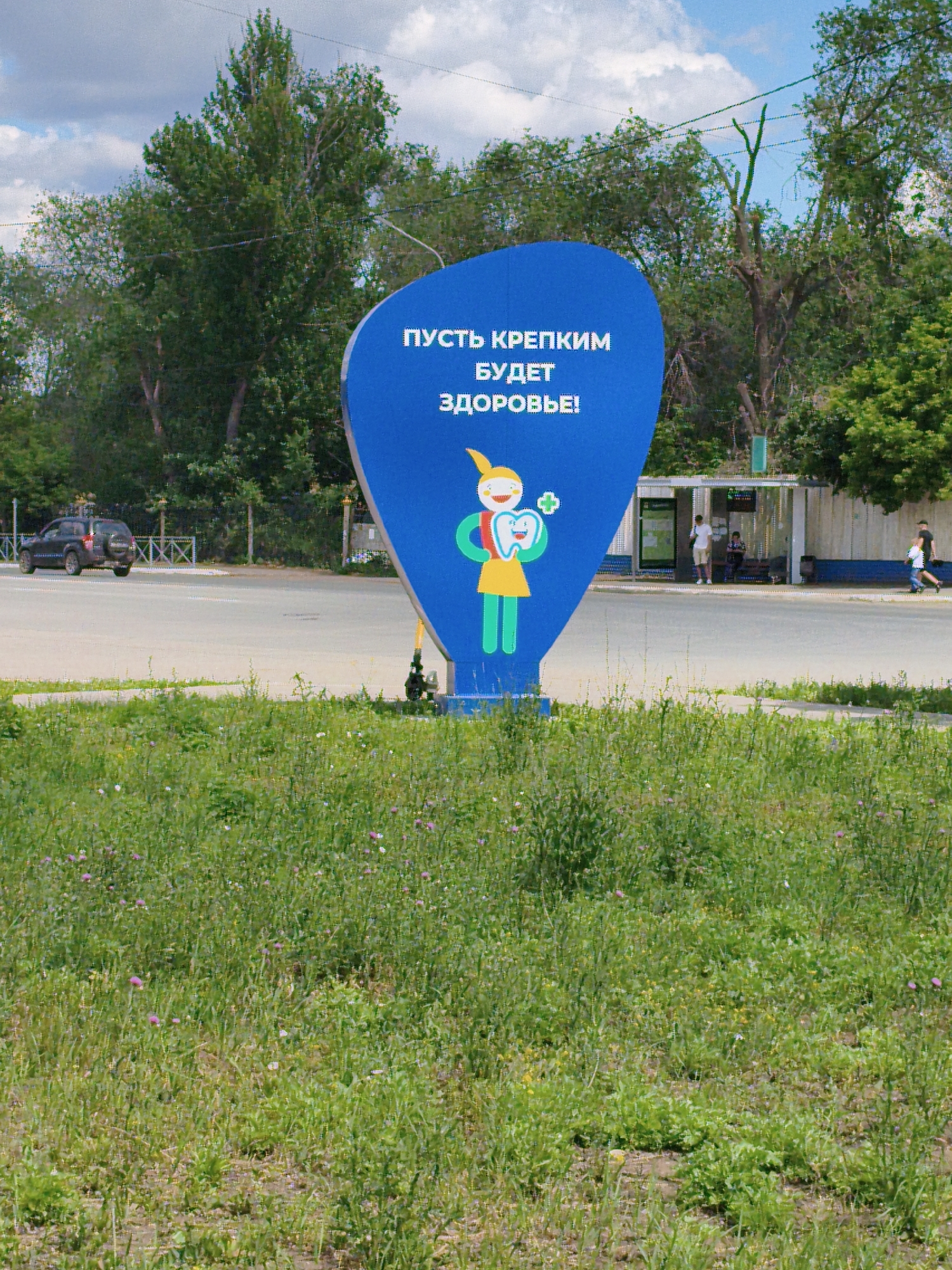 The image shows a blue, speech-bubble-shaped billboard in a public area. The billboard features text in Russian, "ПУСТЬ КРЕПКИМ БУДЕТ ЗДОРОВЬЕ!" which translates to "May your health be strong!". It also depicts a cartoon character of a child holding a large tooth with a green cross symbol. The billboard is located in Orenburg, Russia, likely in a public space near a road and a bus stop. A black SUV is parked on the road, and several people are visible at the bus stop, waiting or walking. The foreground is dominated by lush green grass with scattered wildflowers, suggesting a park or a roadside green space. The background consists of trees and a cloudy blue sky.
