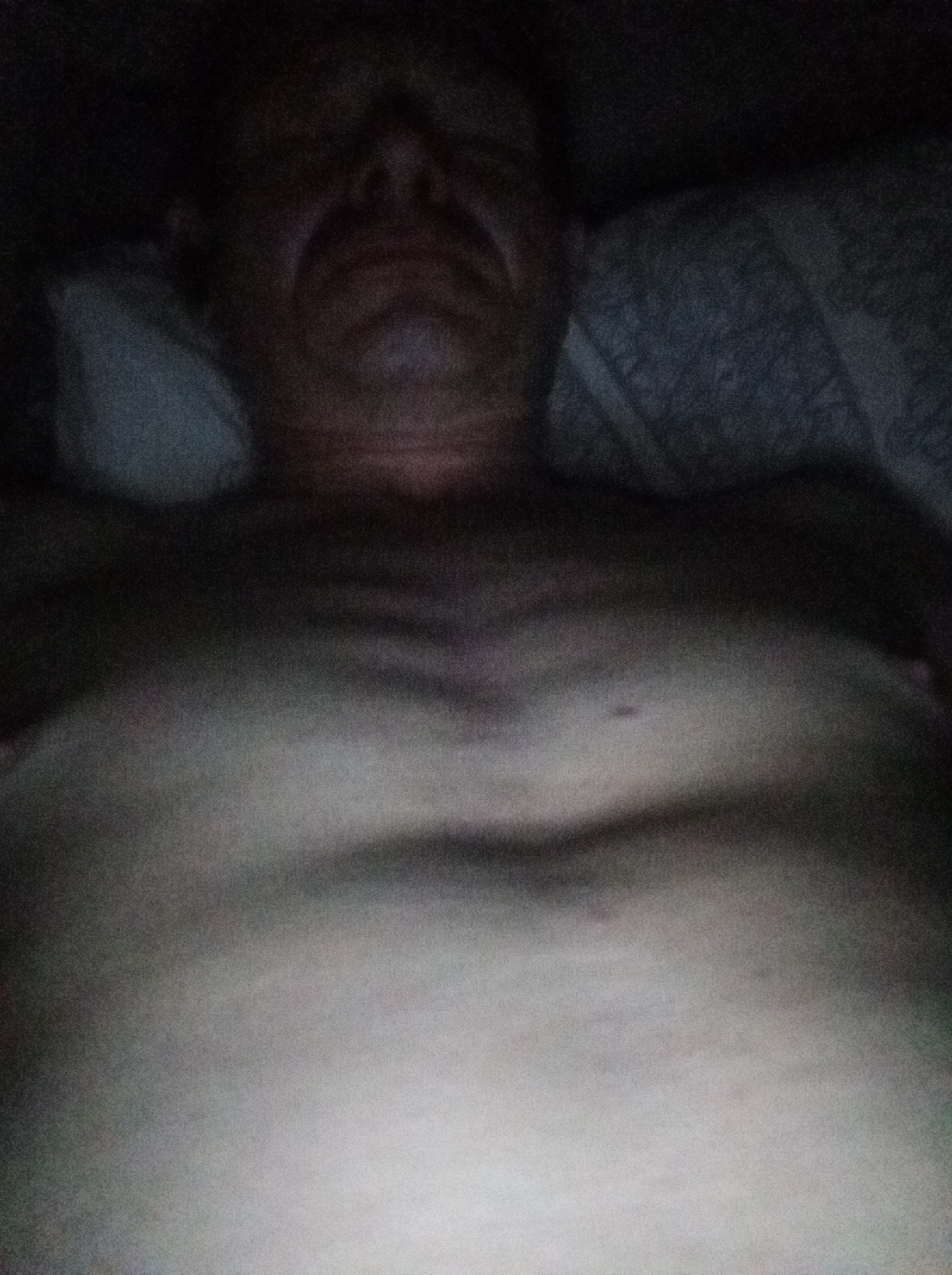 An underexposed, grainy, medium-close-up, eye-level shot depicts the bare torso of a person lying down in bed. The lighting appears to be artificial and dim, creating deep shadows and washing out details. The person's head is visible in the upper portion of the frame, but is largely obscured by shadow. The chest and upper abdomen are visible, with the pectoral muscles and rib cage faintly discernible through the shadows. The background consists of what appears to be a pillow and bedding, also mostly in shadow. The image quality is poor due to low light conditions and noise, making it difficult to discern specific features or textures. The scene appears to be an indoor domestic setting, likely a bedroom. There is no indication of the location being in Tapa, Estonia.