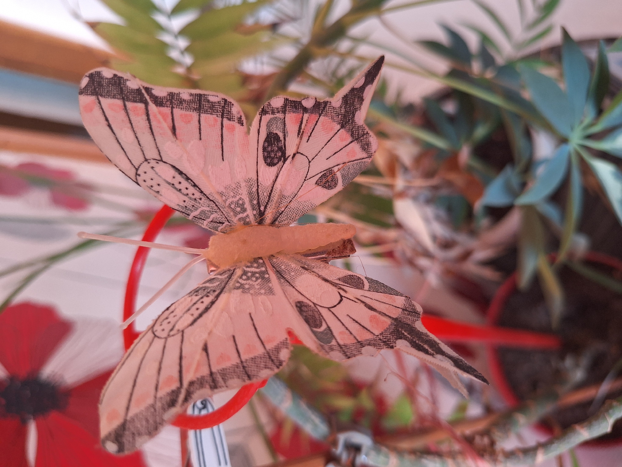 A close-up, shallow depth of field photograph captures a decorative pink and black butterfly attached to a red object, possibly a plant support. The butterfly's wings are spread open, displaying intricate vein patterns and delicate pink speckles. The butterfly's body is a light tan color and appears fuzzy. In the background, out-of-focus green foliage and parts of a red poppy are visible, suggesting an indoor setting with plants, possibly near a window. The overall impression is of a whimsical, decorative element within a natural or simulated natural environment. The image does not provide any identifiable geographic markers or context to place it in Varna, Bulgaria.