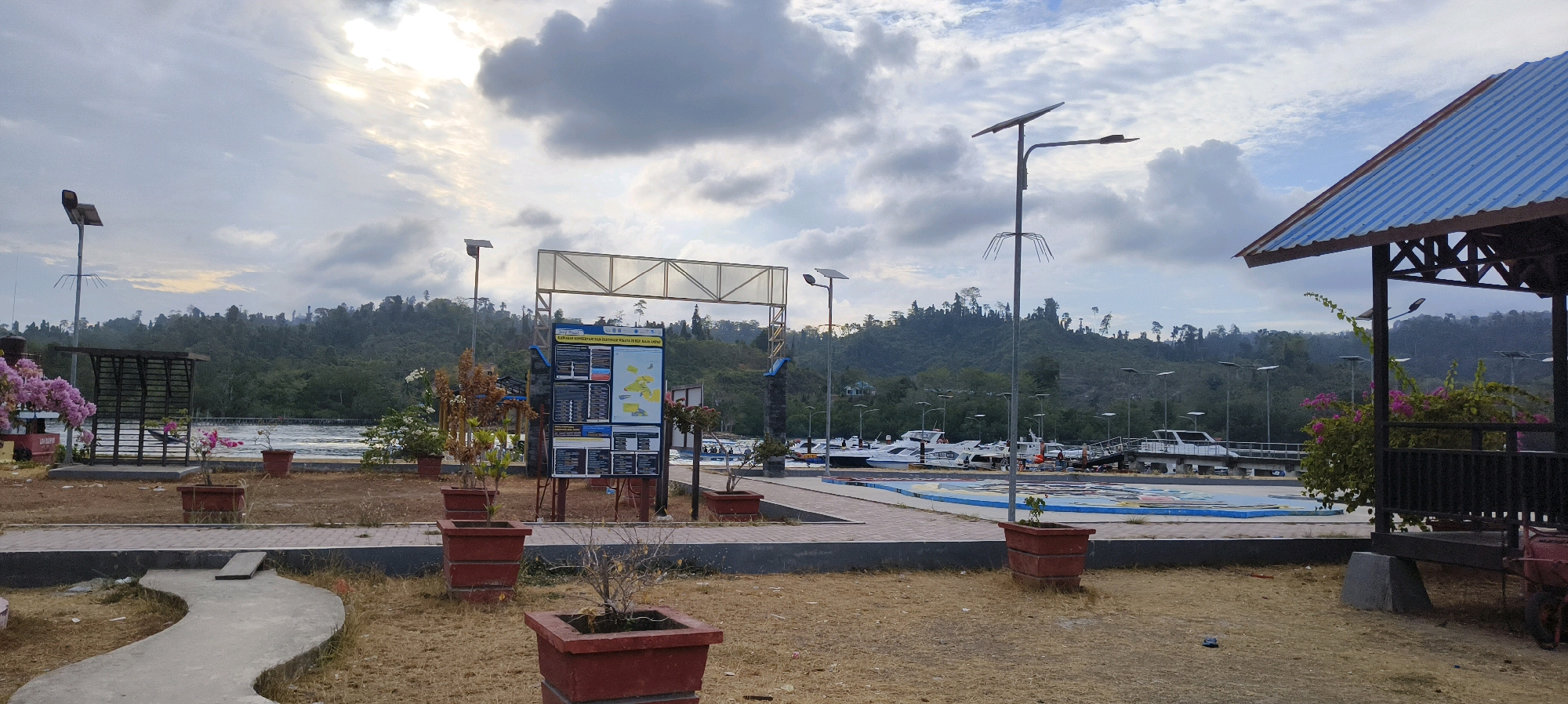 The image depicts the waterfront of Waisai, Indonesia, likely a harbor or pier area. The foreground shows dry, yellowish grass and red rectangular planters. A winding grey concrete path is visible in the lower left corner. In the midground, there is a paved walkway with a central informational display board, supported by a metal frame. To the left of the display, a shaded structure with a dark roof and open sides is present, with pink flowering plants nearby. Further to the left, another shaded structure with a slatted roof is visible.
The main focus of the midground and background is a body of water with several white motorboats docked. A pier extends into the water where some of these boats are moored. In the background, a lush green, forested hillside rises, with a hazy sky overhead. Several lampposts, some equipped with solar panels, are scattered throughout the scene. A gazebo-like structure with a blue corrugated metal roof is visible on the right side of the image, with dark railings and a concrete base. The lighting suggests it is either early morning or late afternoon.
