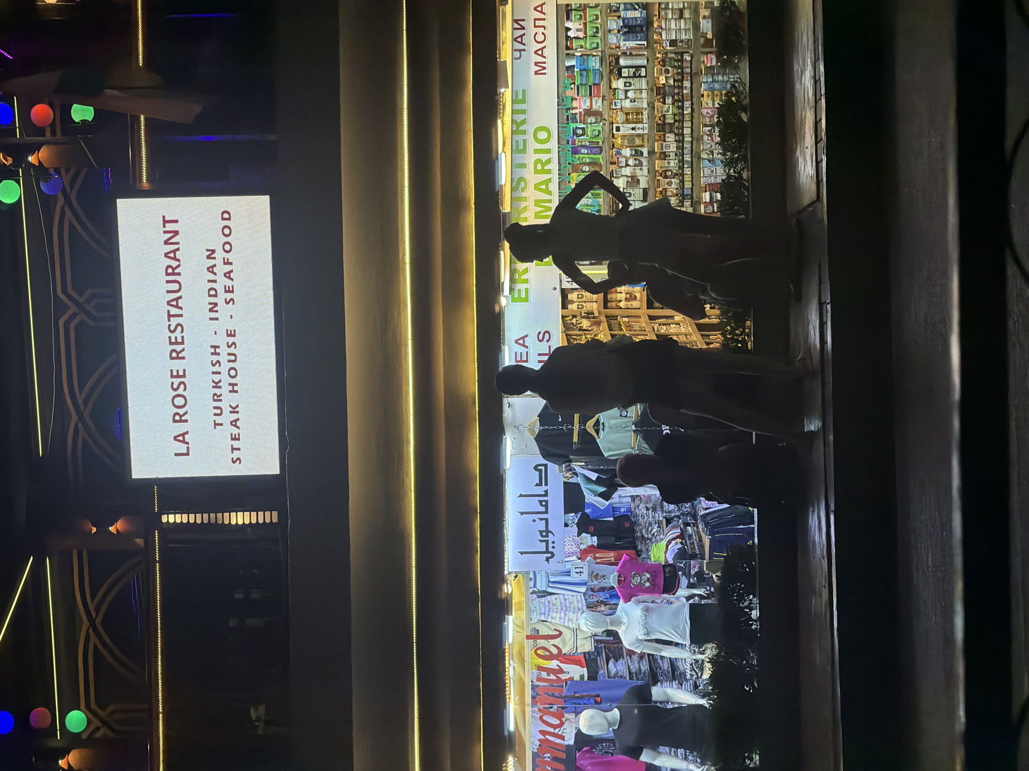 The image depicts the exterior of a storefront in Sharm El Sheikh, Egypt. A large illuminated sign above the storefront reads "LA ROSE RESTAURANT TURKISH - INDIAN STEAK HOUSE - SEAFOOD." Below this, the storefront displays clothing on mannequins and racks, with Arabic text visible on signage. To the right of the clothing display, a shop selling various bottles and products is visible through the glass. Silhouettes of individuals are seen in the foreground, appearing to be patrons or passersby observing the storefronts. The lighting suggests it is nighttime, with colorful lights and illuminated signage creating a vibrant atmosphere.