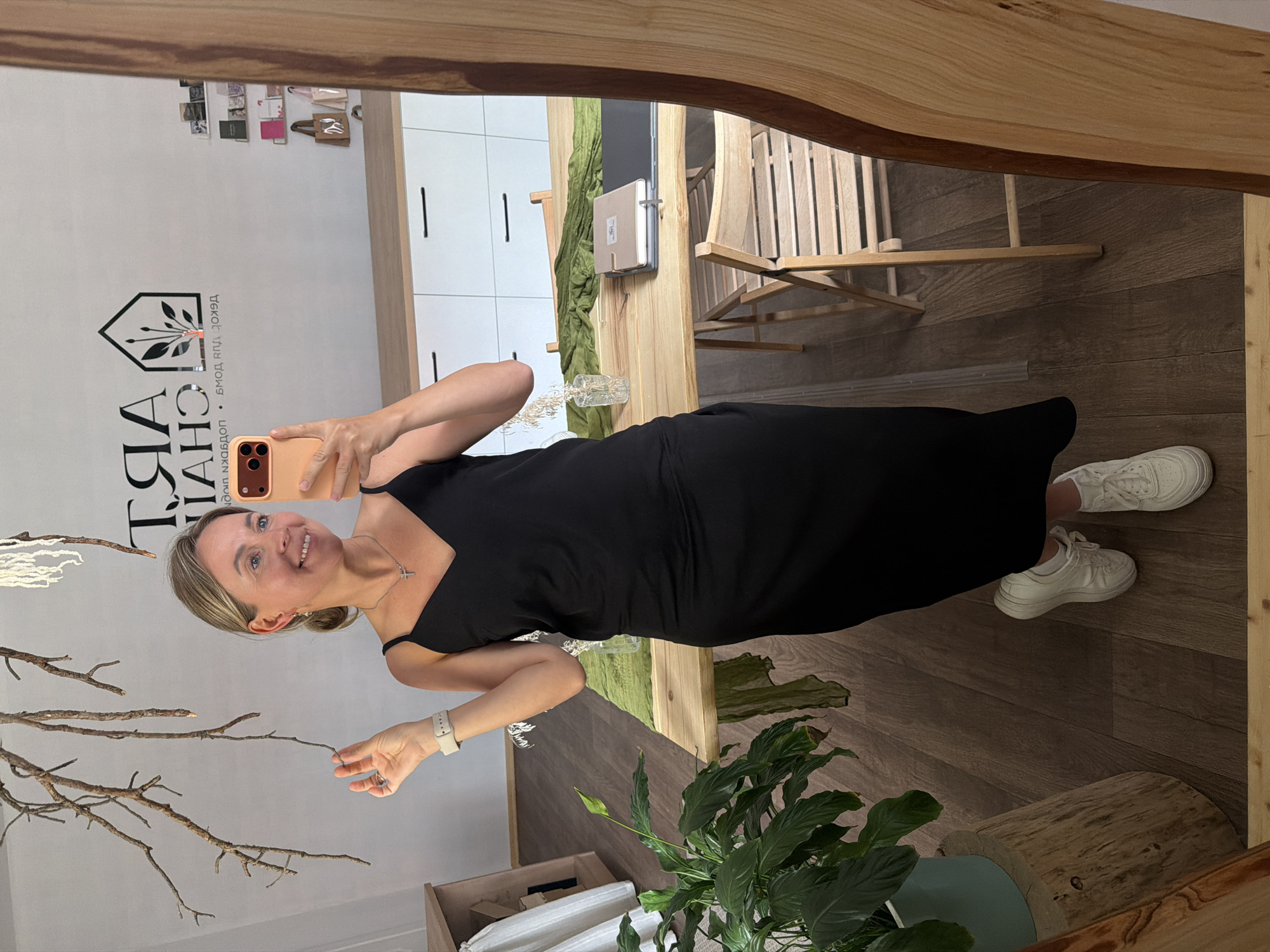 The image shows a woman standing in what appears to be a shop or studio. She is wearing a black slip dress, white sneakers, and a silver necklace. She is holding a smartphone in her right hand and looking into a mirror. The background shows a wooden table with some items on it, white cabinets, and a logo on the wall that reads "ART CHAI". The floor is made of wood. The overall scene suggests an interior space, possibly a retail or creative environment. The logo "ART CHAI" is a strong indicator that this location is in the city of Chaikovsky, Russia, as "Chai" is a Russian word and likely refers to the city's name.