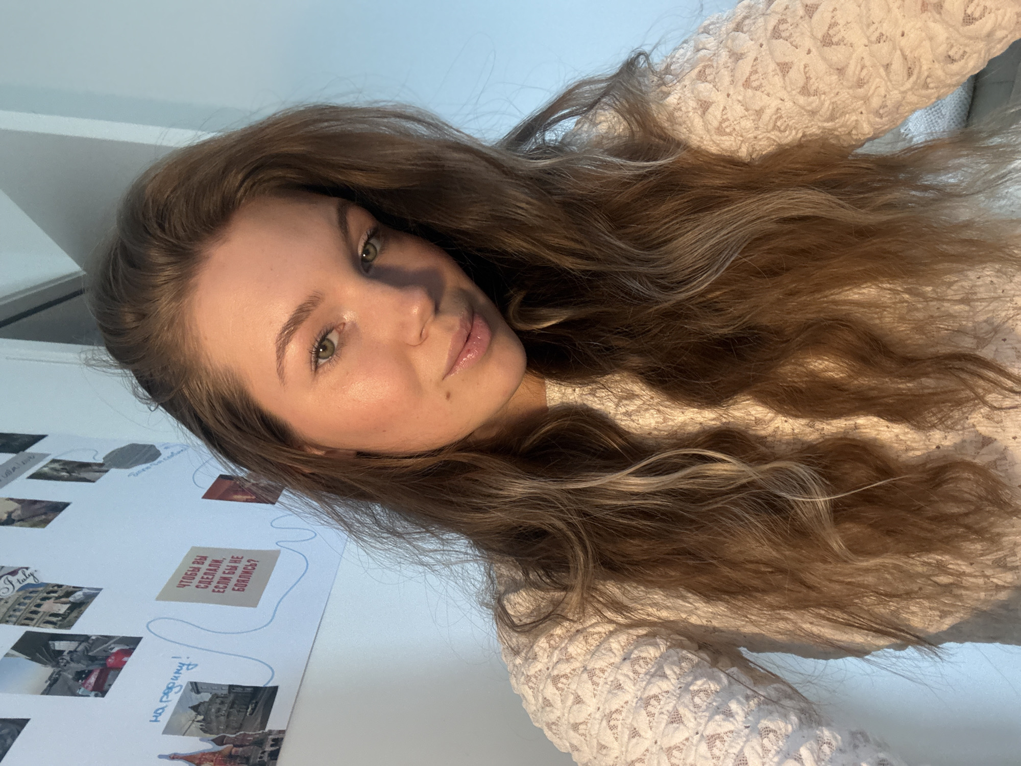A young woman with long, wavy brown hair is posing for a selfie. She is wearing a white lace top. In the background, a white wall is visible with a collage of photos and text in Russian. The location cannot be determined from the provided information.