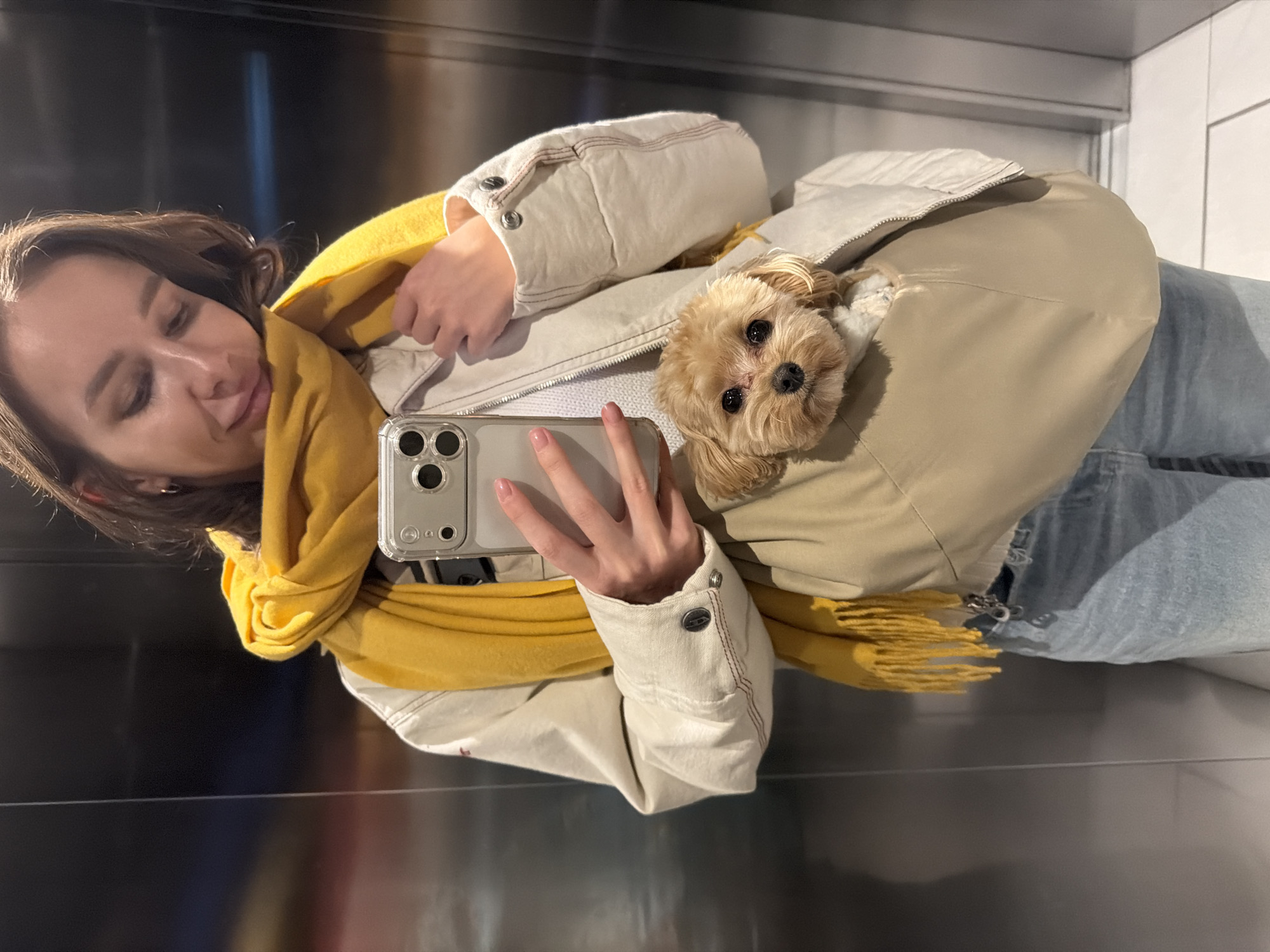 A young woman takes a selfie indoors. She is wearing a yellow scarf and a light-colored jacket with a dog inside. She is holding an iPhone. The location is most likely in Moscow, Russia.