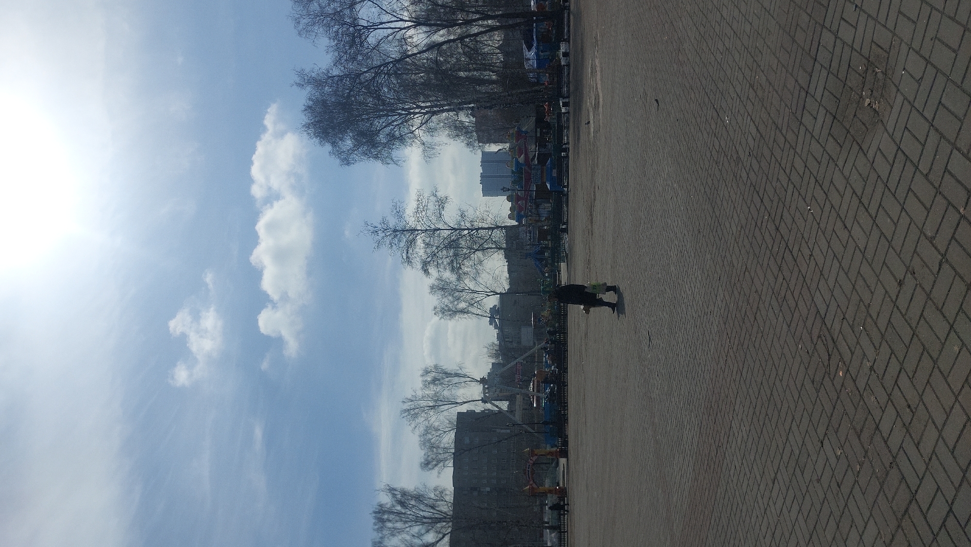 The image depicts a sunny, likely early spring day in a park in Novosibirsk, Russia. The sky is blue with scattered cumulus clouds and a bright sun, partially obscured. The foreground is a paved area with a herringbone brick pattern. A person, possibly carrying a bag, walks across the paved area. In the background, bare trees line the park. Behind the trees, there are buildings, including some amusement park rides.