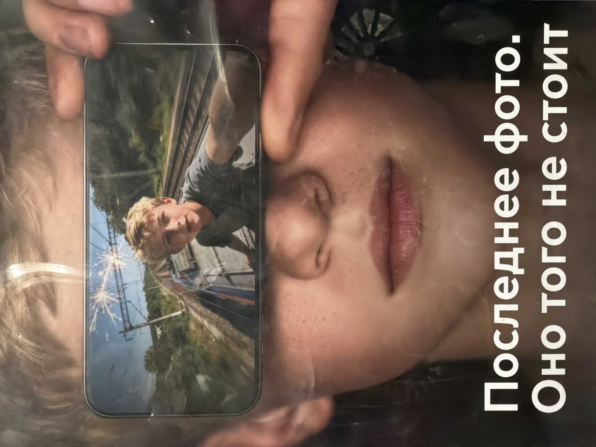 A photo shows a person holding a smartphone in front of another person's face. The smartphone displays an image of a person standing near a train track. Sparks appear to be flying. Text in Cyrillic script is visible to the right of the face. The background is indistinct. The location is not identifiable as Moscow, Russia.