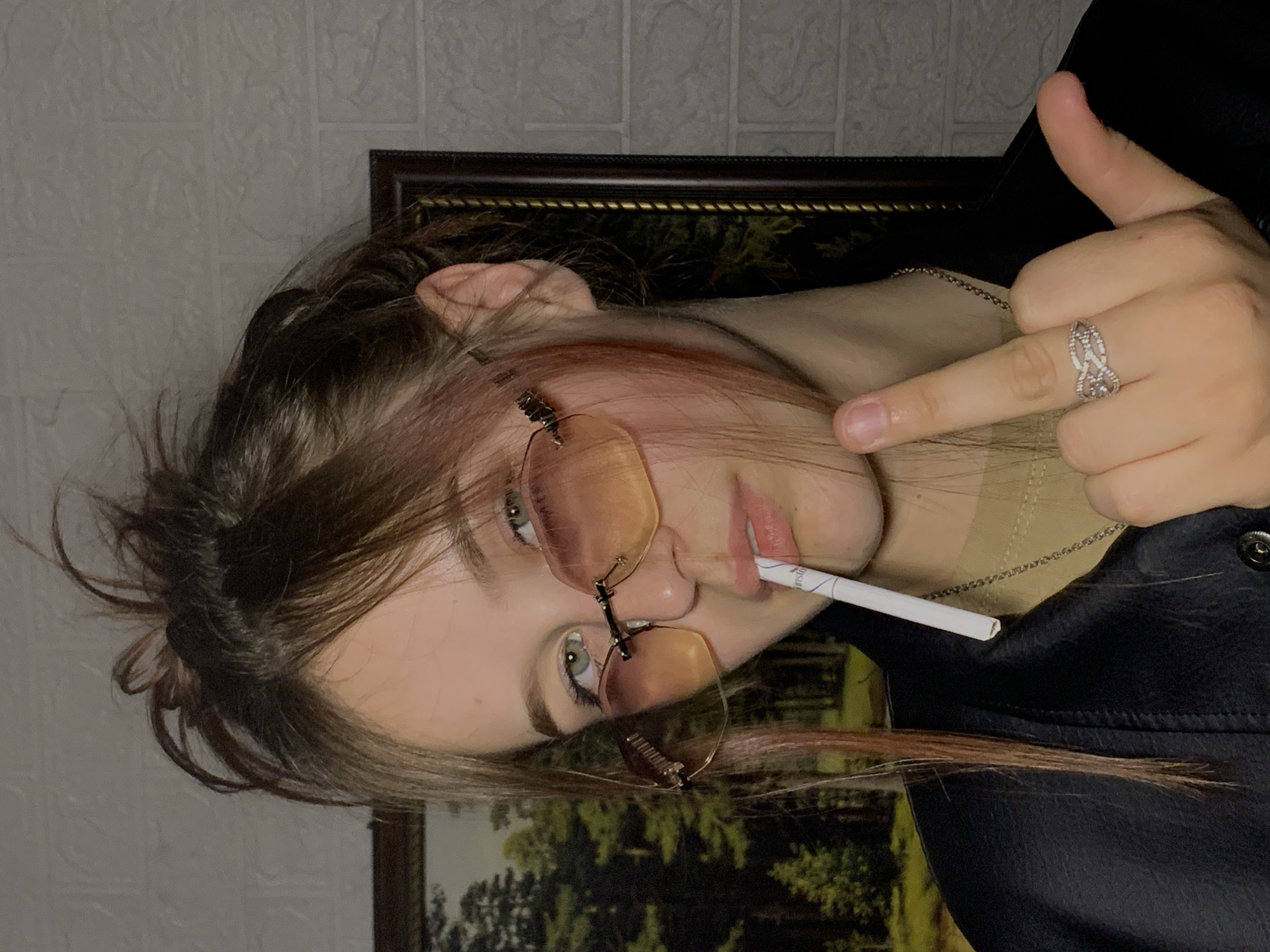 A female individual with fair skin, green eyes, and brown hair wearing gold-tinted rimless glasses is shown holding a lit cigarette in her mouth and extending her middle finger toward the viewer. She is wearing a silver ring on her middle finger and a silver necklace. She is dressed in a black garment. The background consists of a brick-patterned wall and a framed painting with visible green and brown hues. The scene appears to be indoors. There is no information on Фоки, Russia, in this image.