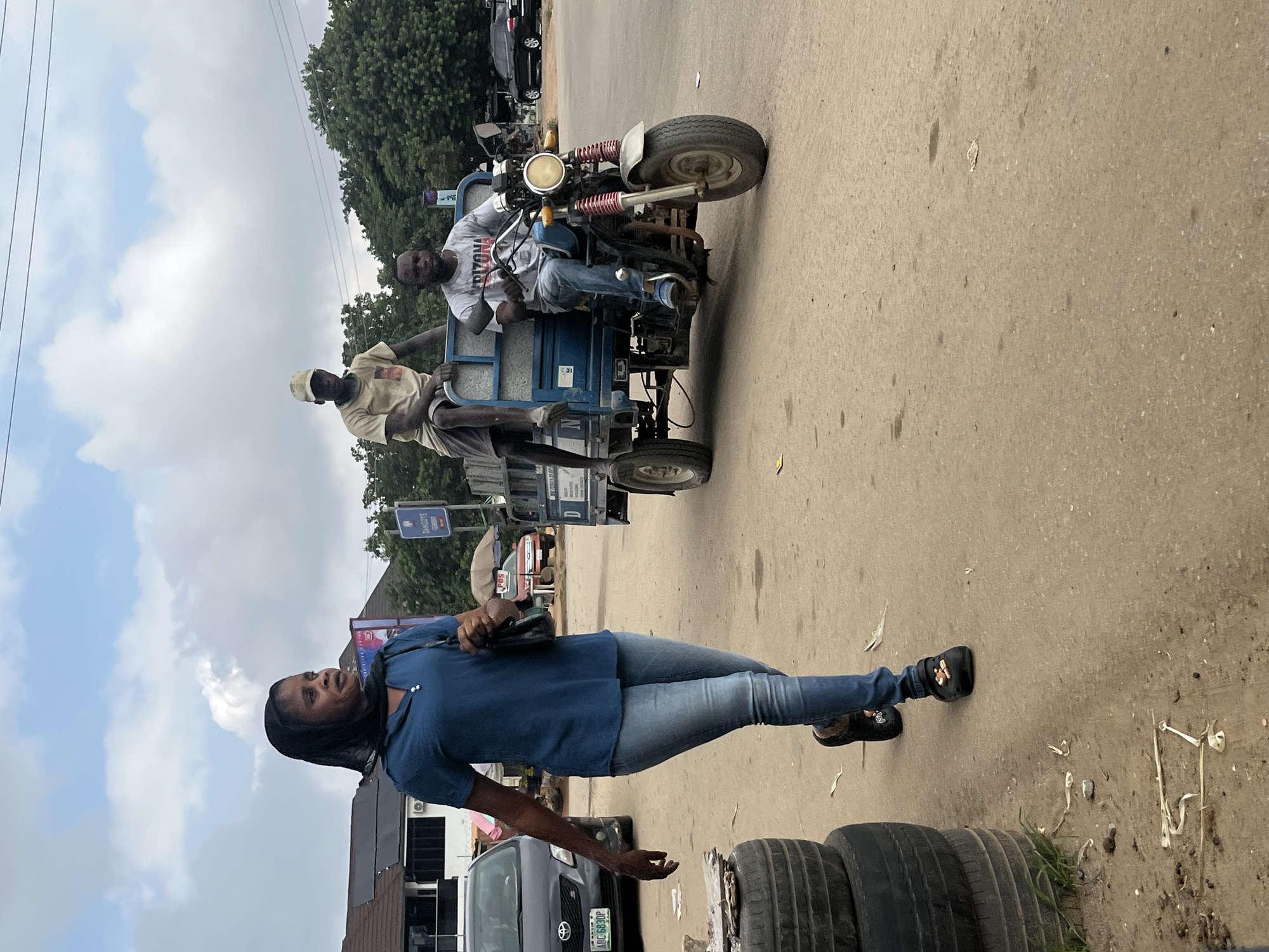The image shows a street scene in Abuja, Nigeria, with several people and a three-wheeled vehicle. A woman in a blue collared shirt and blue jeans stands on the right side of the frame, looking to her left. She is wearing black Crocs with a colorful design on them. In front of her, two tires are stacked, and to her left, part of a grey car with a green and white license plate is visible.
Behind the woman, a three-wheeled motorcycle-like vehicle is on the road. Two men are seated on it. The driver, wearing a grey t-shirt with "ARIZONA" printed on it and blue jeans, is at the front. Behind him, another man in a yellow t-shirt and a light-colored cap is also seated. The vehicle has a blue cargo bed. In the background, trees, a cloudy sky, and other vehicles suggest an outdoor, daytime setting. The overall impression is a typical street scene in a busy urban environment.